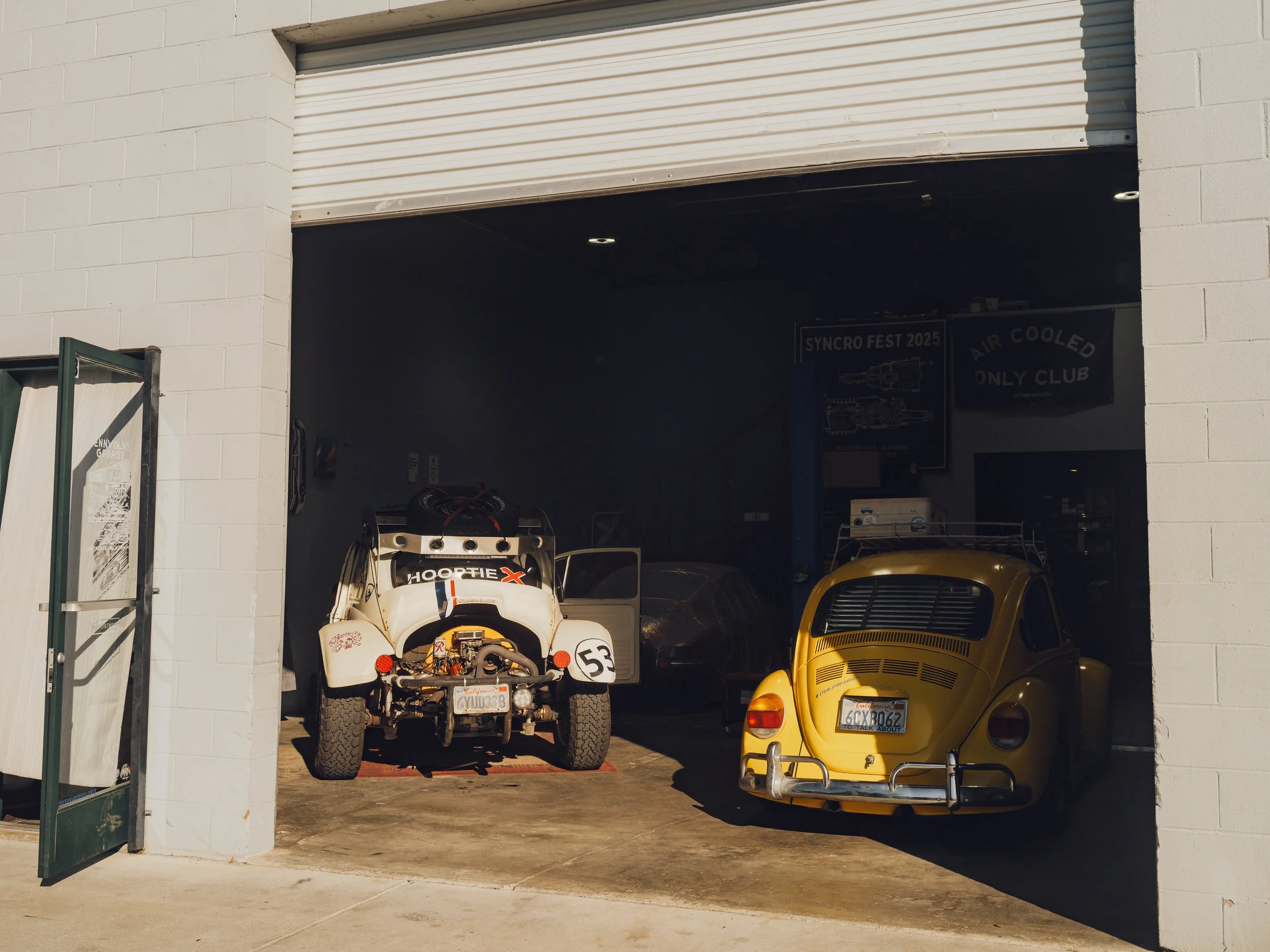 A garage with two cars visible: a white off-road vehicle with a black roof rack and a yellow classic Volkswagen Beetle with red and orange tail lights.