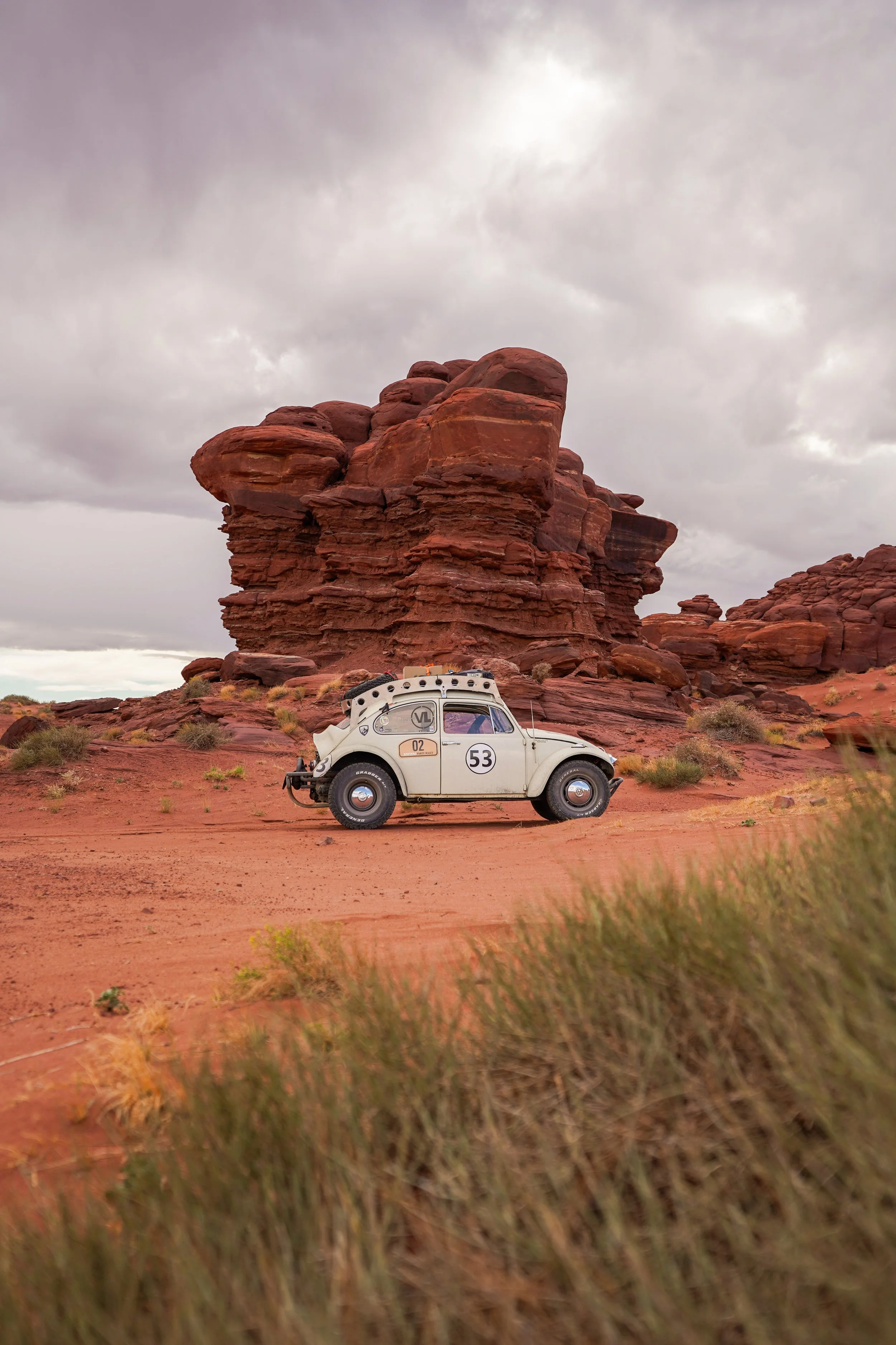 A vintage white Volkswagen Beetle car parked on desert terrain with reddish rock formations and cloudy sky in the background.