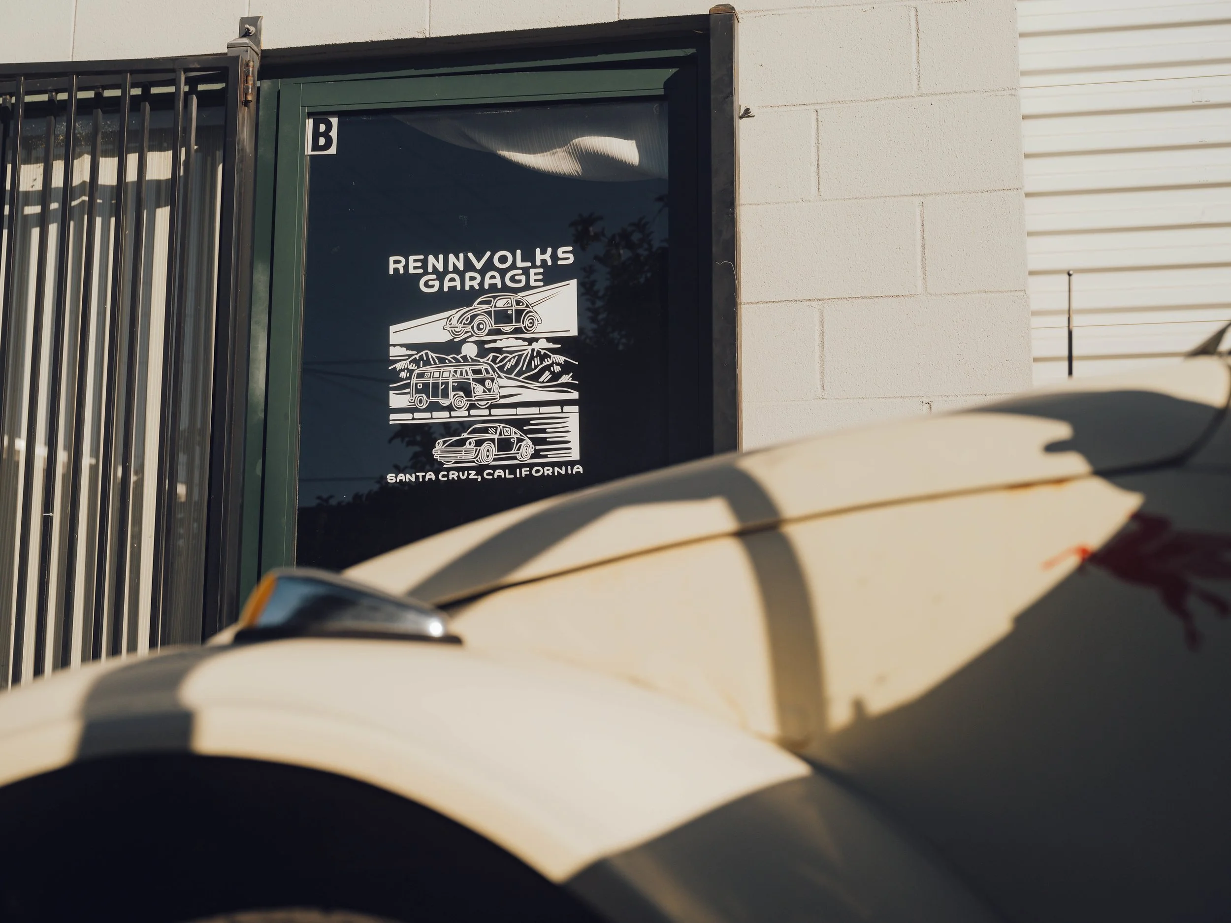 Close-up of the front of a white car parked outside Rennvolks Garage in Santa Cruz, California, with the garage's sign visible in the background.