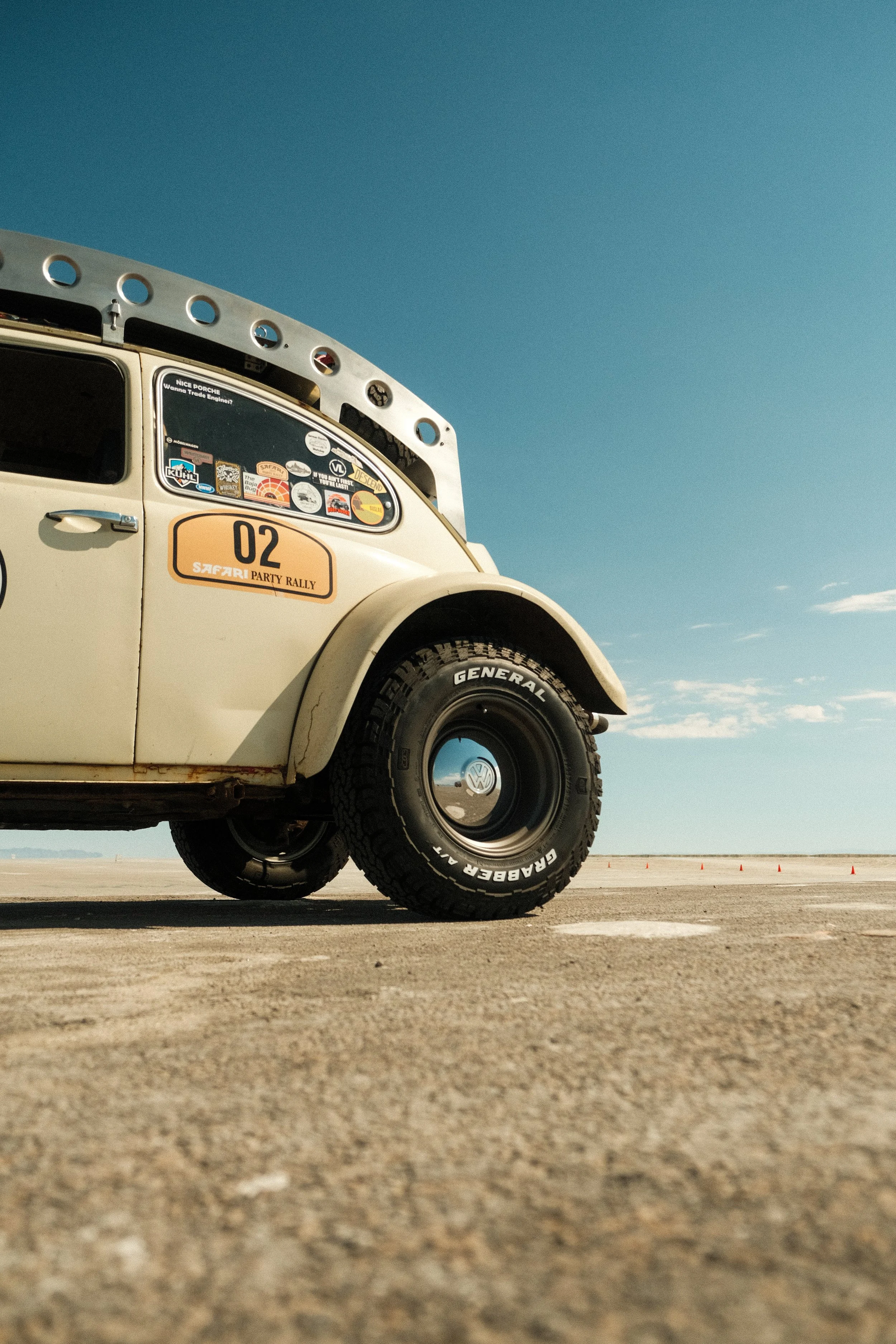 Close-up of a vintage Volkswagen Beetle with stickers on the rear window, parked on a flat, open surface under a blue sky with a few clouds.
