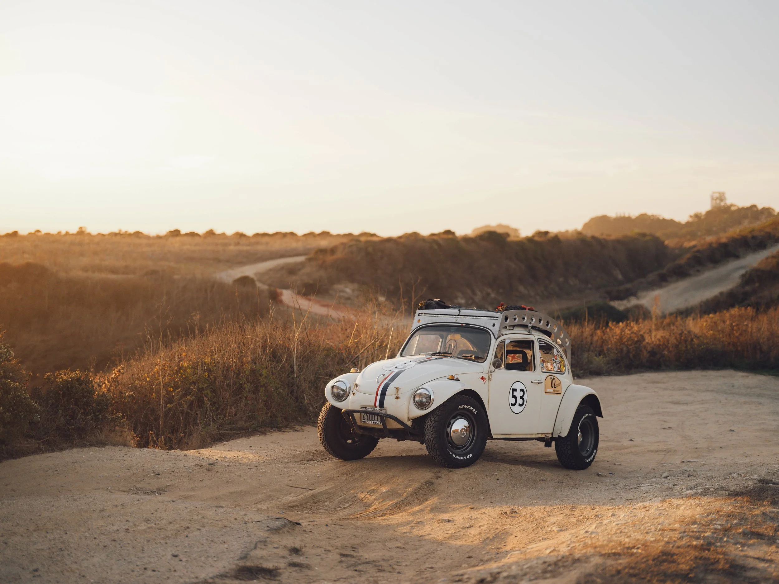 A small vintage white off-road vehicle with racing decals parked on a dirt road in a desert landscape at sunset.