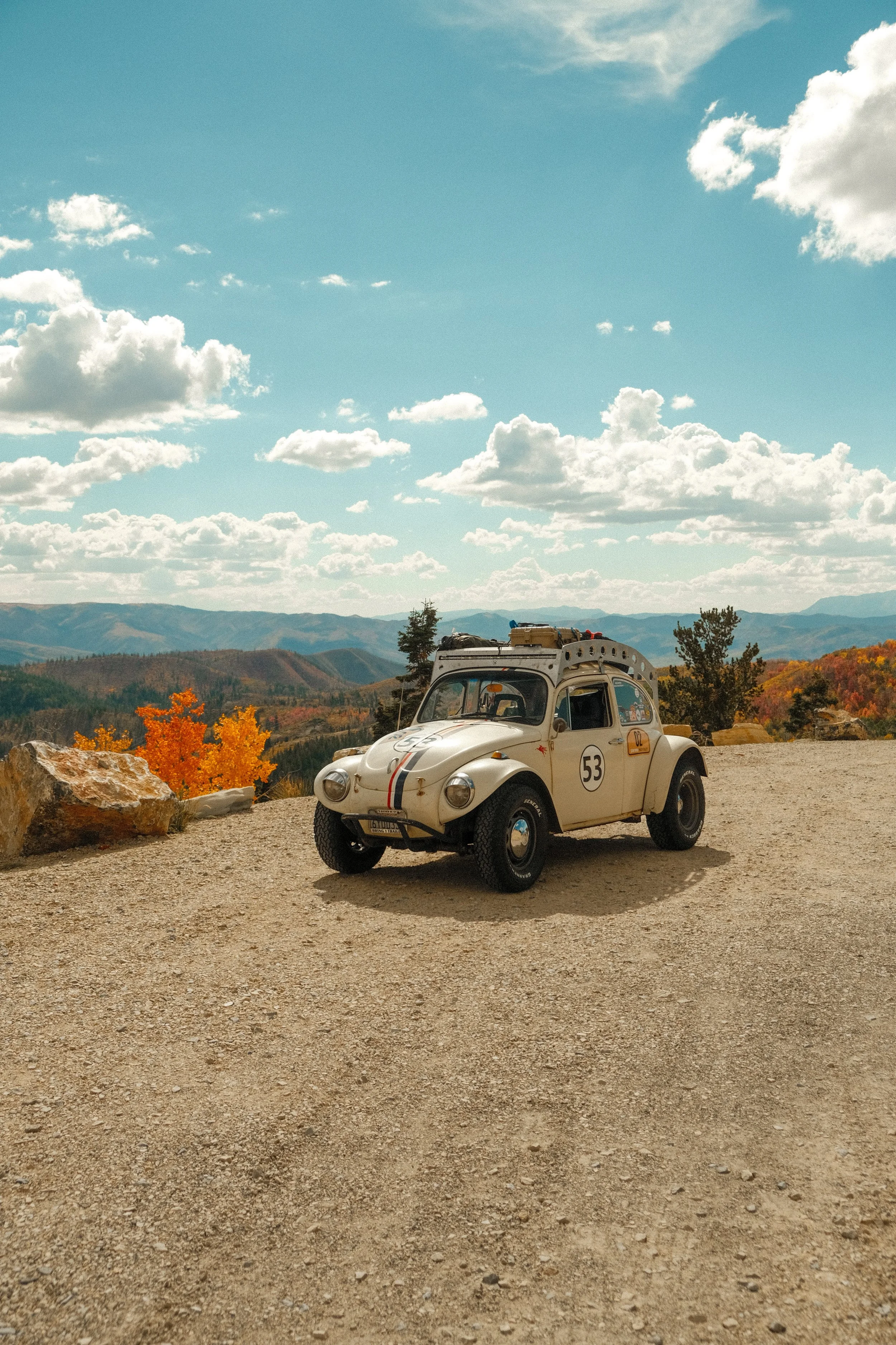 A vintage beige Citroën 2CV car with racing stripes and racing numbers parked on a gravel overlook with autumn trees and mountain range in the background under a partly cloudy sky.