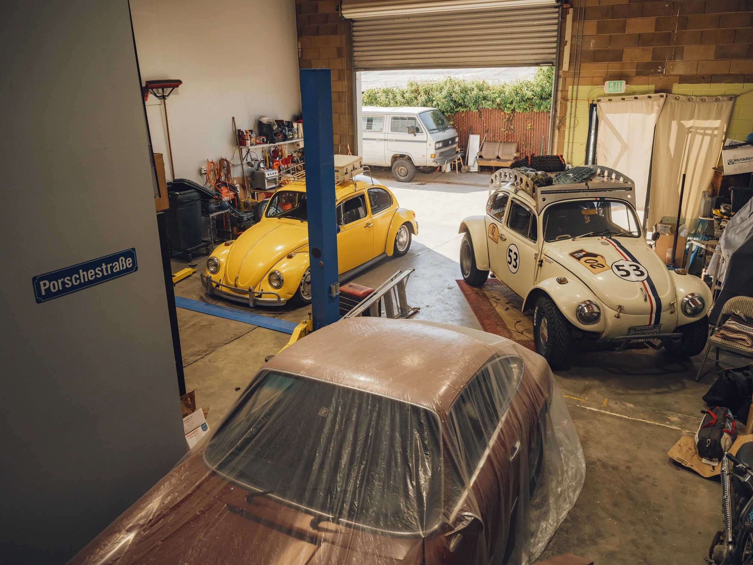 A garage with a yellow classic Volkswagen Beetle, a white racing Volkswagen Beetle with racing decals and a number, and a brown car covered with a plastic sheet. There are tools and equipment on the shelves and a partly open garage door showing an ou