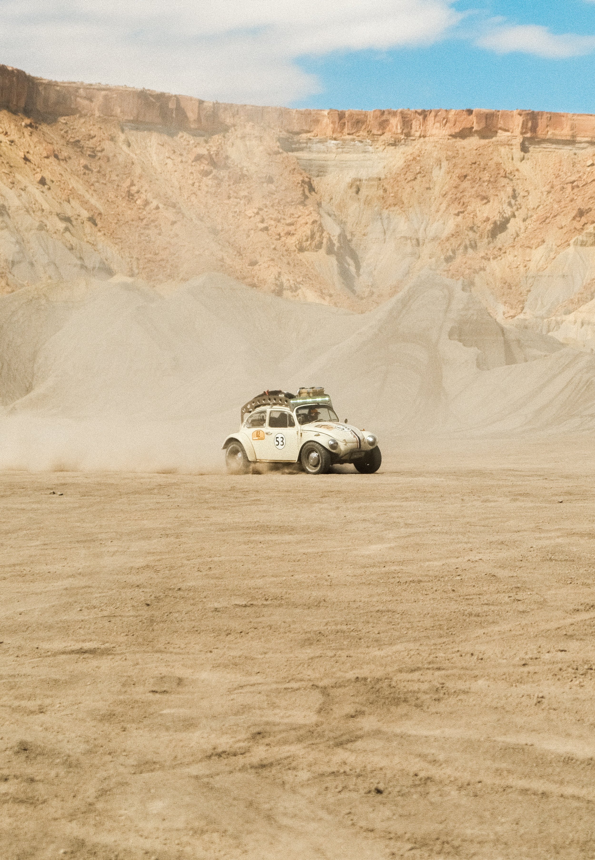 A vintage cream-colored car driving across a dusty desert landscape with a rugged canyon in the background.