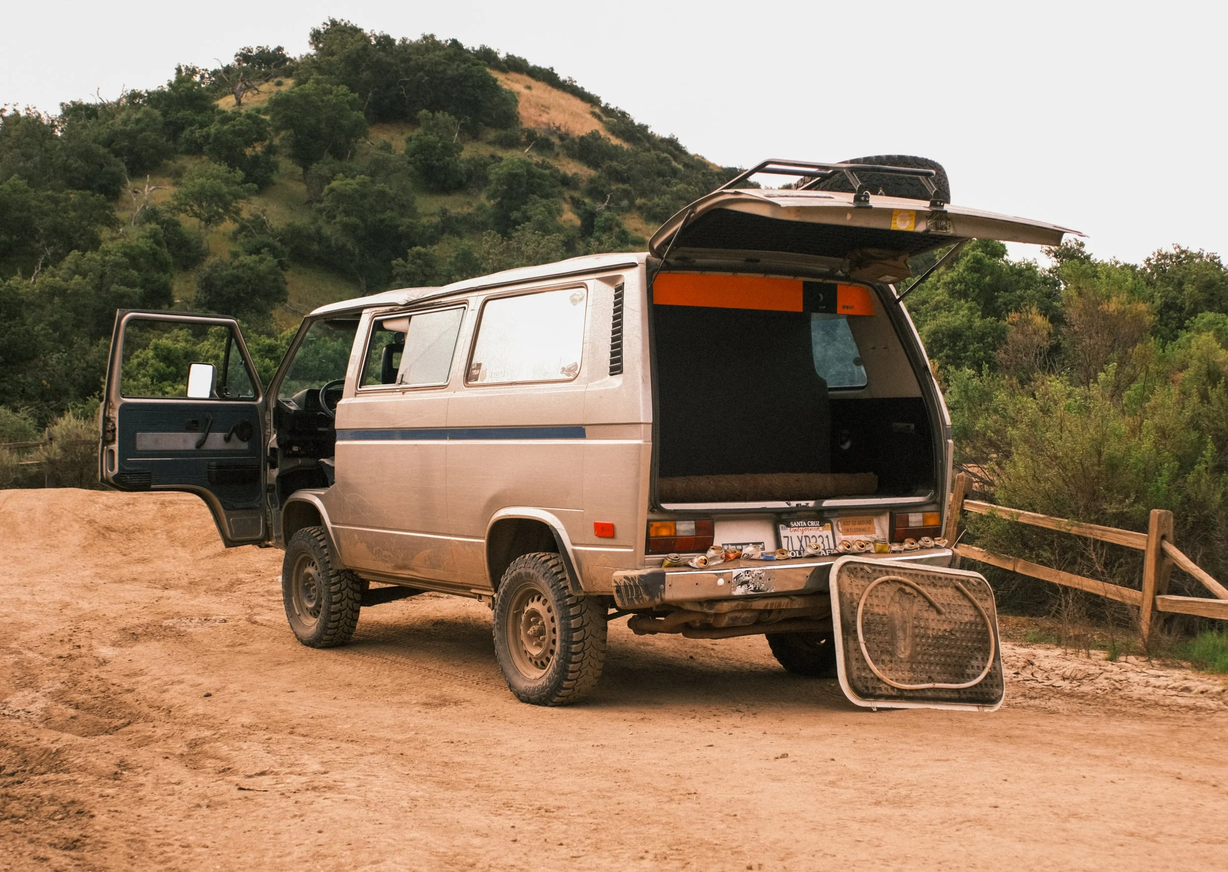 A vintage silver van with an open rear hatch and a grille on the ground behind it, parked on a dirt road with green hills and trees in the background.