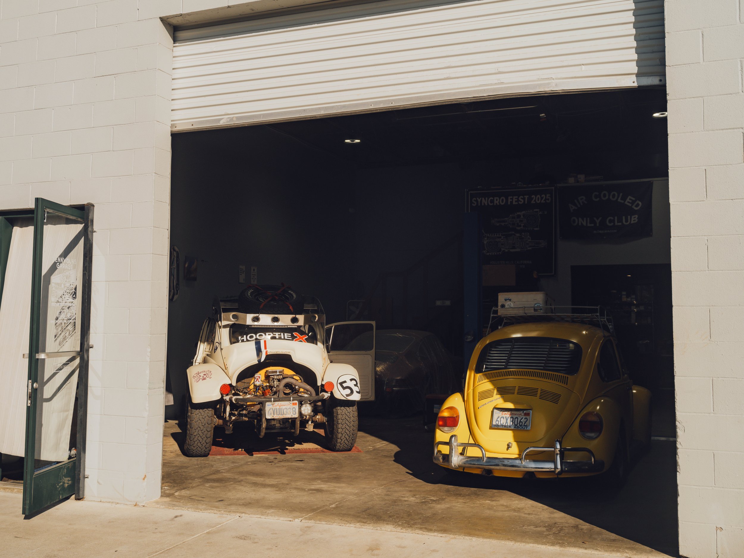 A garage door open to reveal a white racing car with the number 53 and a yellow vintage Volkswagen Beetle parked inside.
