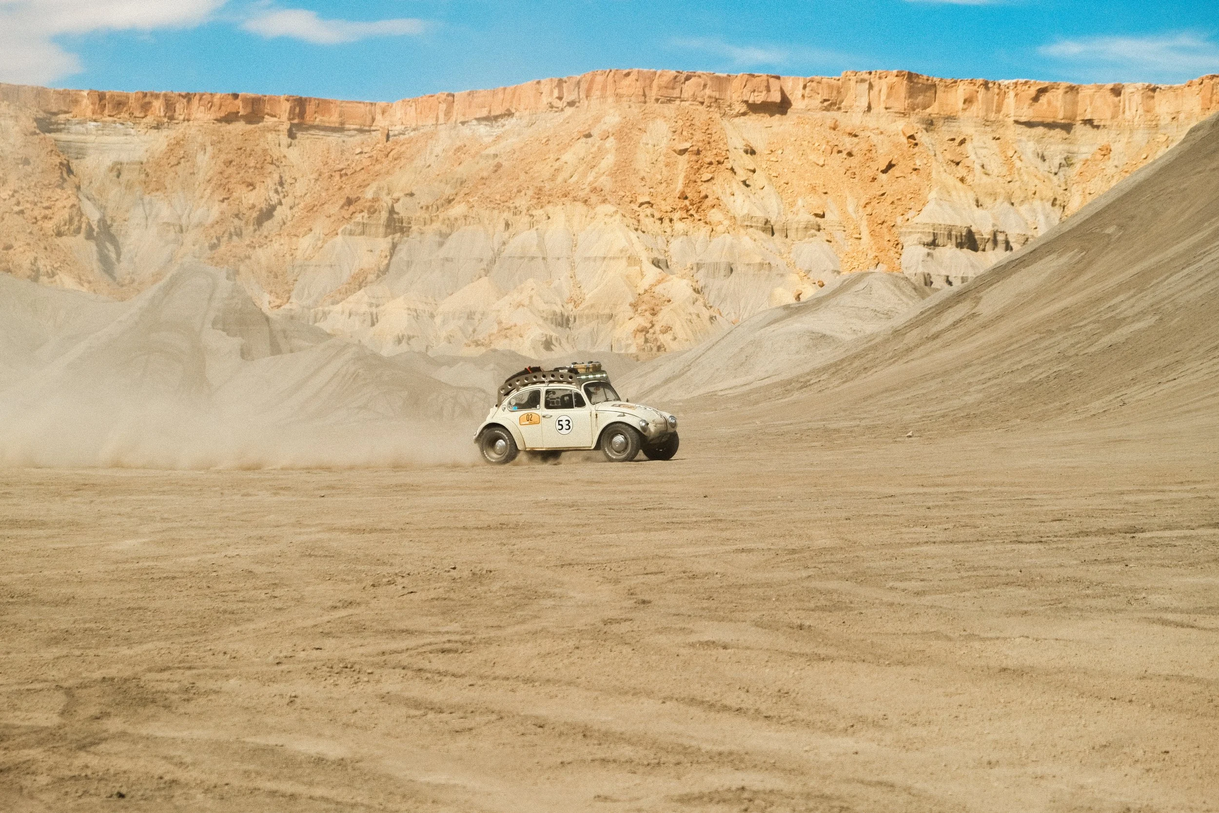 A vintage white car with a roof rack driving across a desert landscape surrounded by mountains with dust clouds behind it.