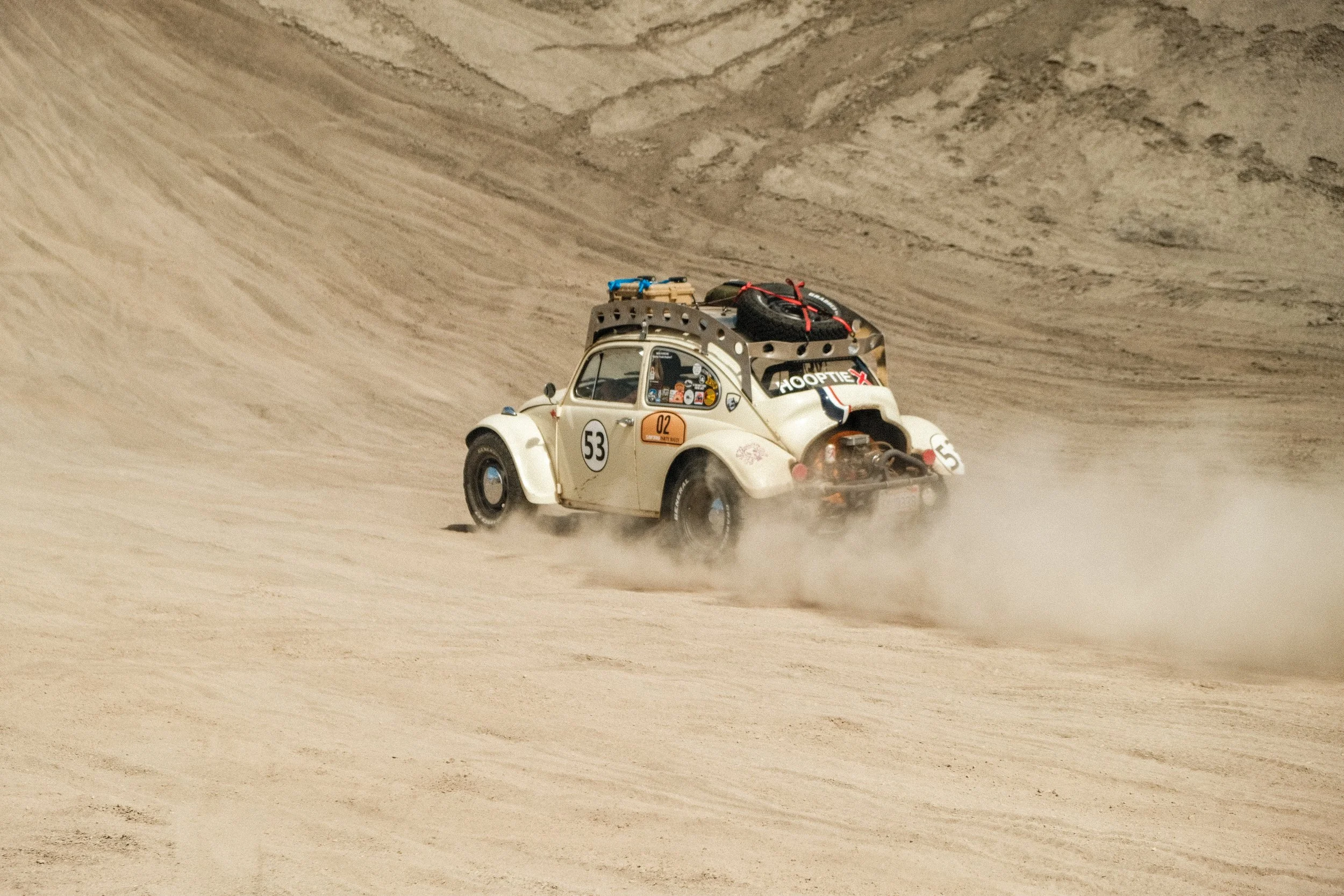A small vintage-style off-road vehicle driving on sandy terrain, kicking up dust with a large hill or dune in the background.
