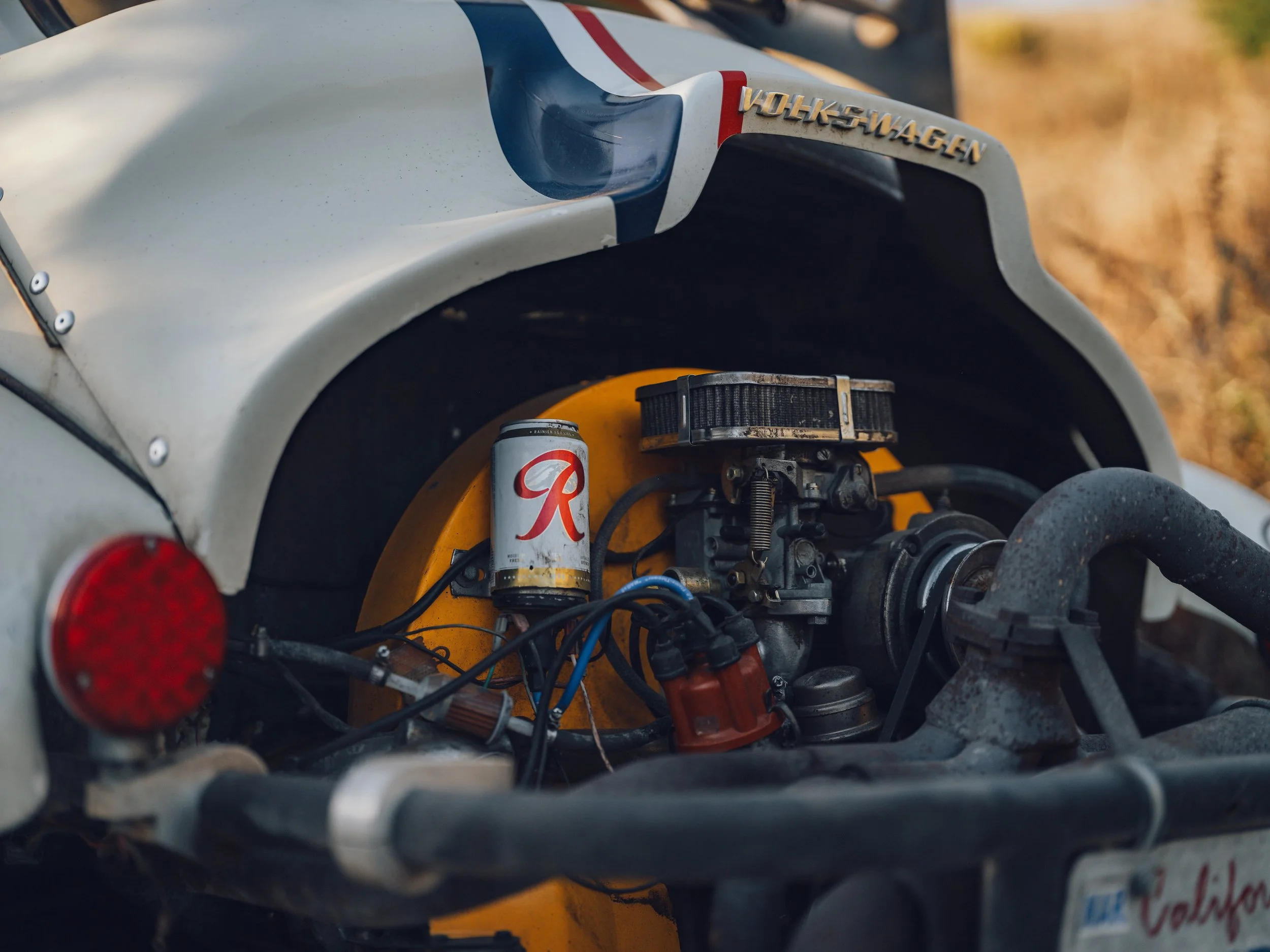 Close-up of a vintage Volkswagen race car engine with visible parts including a Volkswagen badge, a can of Red Bull, and various engine components, set against a blurred outdoor background.