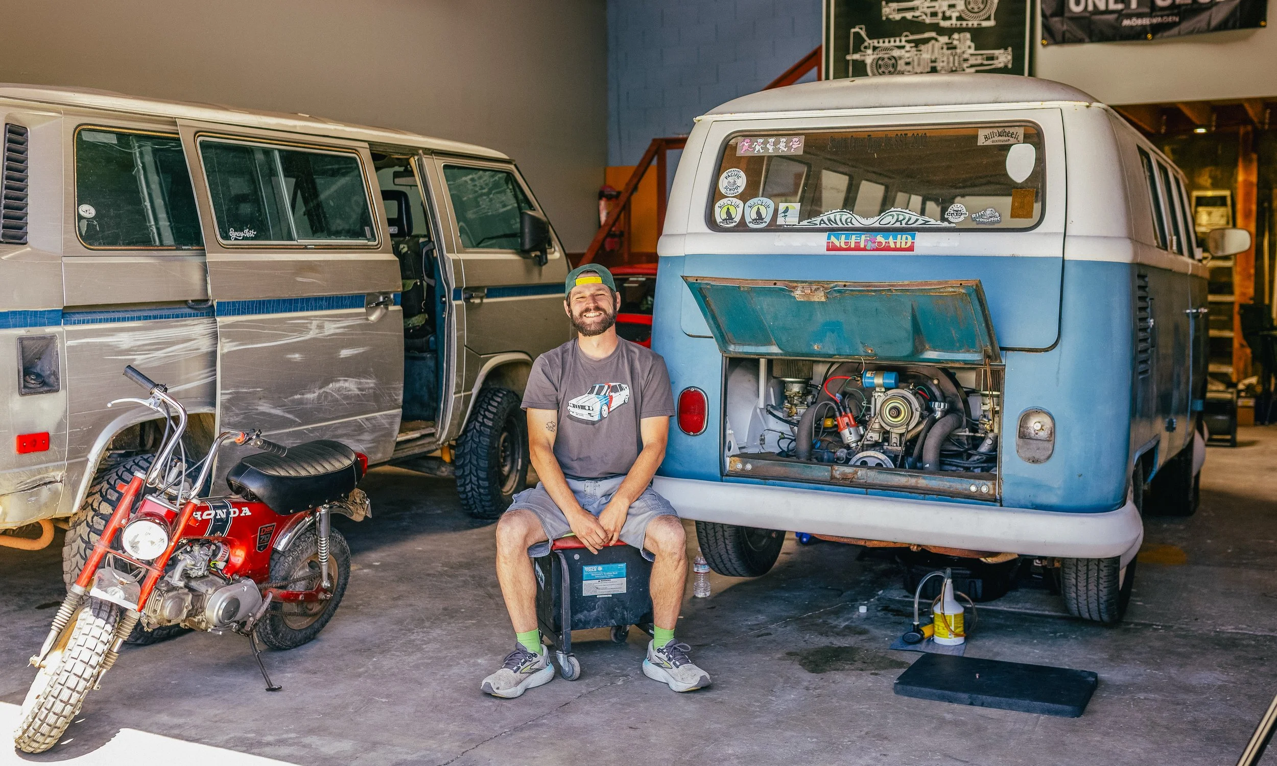A man sitting on a toolbox with a vintage blue camper van and a red Honda motorcycle in a garage.