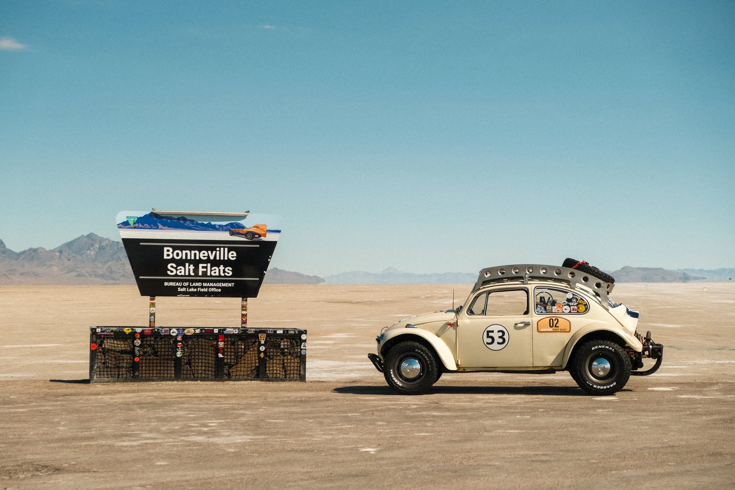 A vintage beige Volkswagen Beetle car with racing stickers parked on a salt flat, with a sign that reads 'Bonneville Salt Flats' in the background.