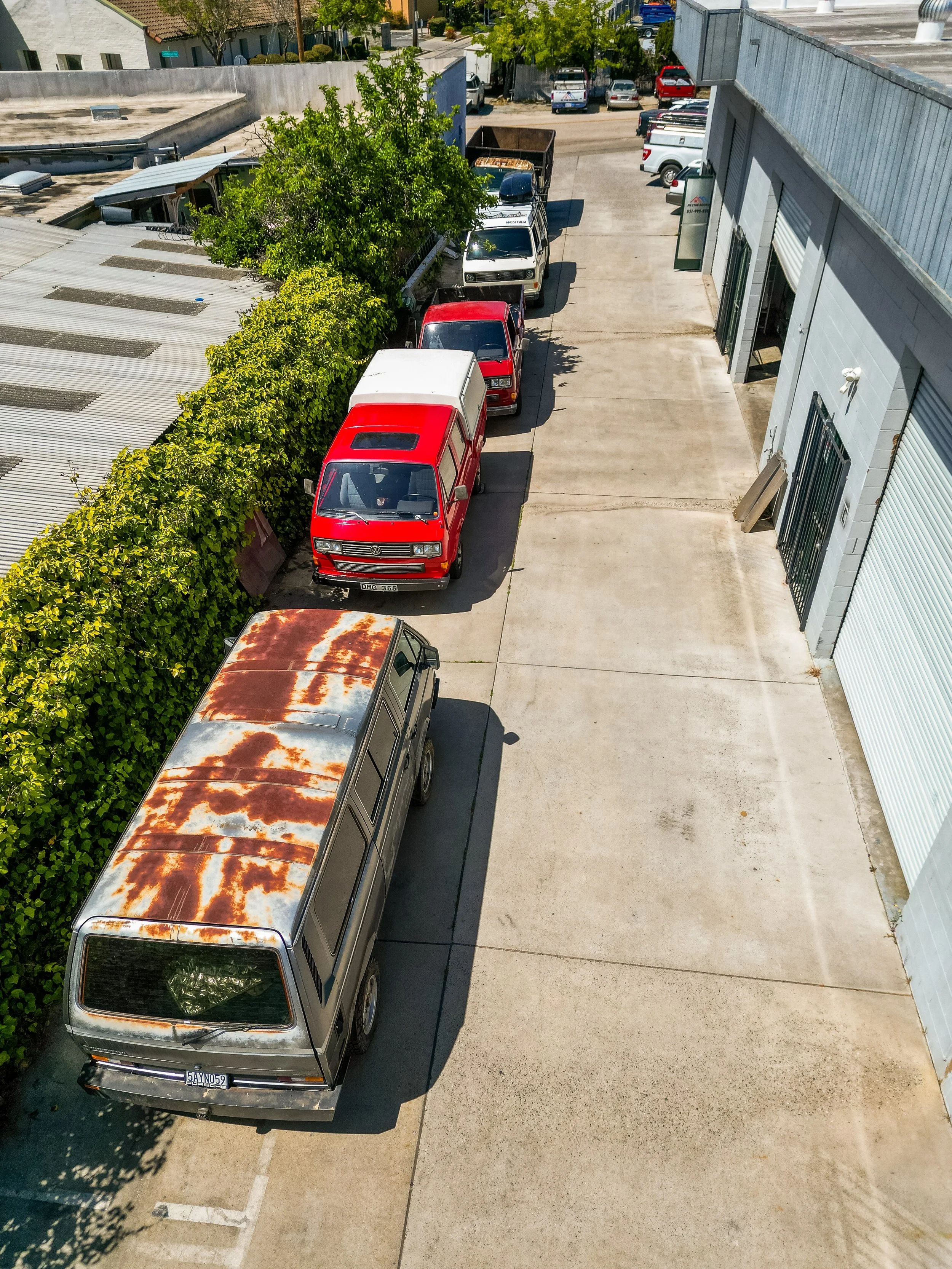 A parking area with several parked vehicles, including a rusted van, a red van, a white van, and a black truck. There is a green hedge on the left and a large gray building with roll-up doors on the right. In the background, there is a sidewalk with 