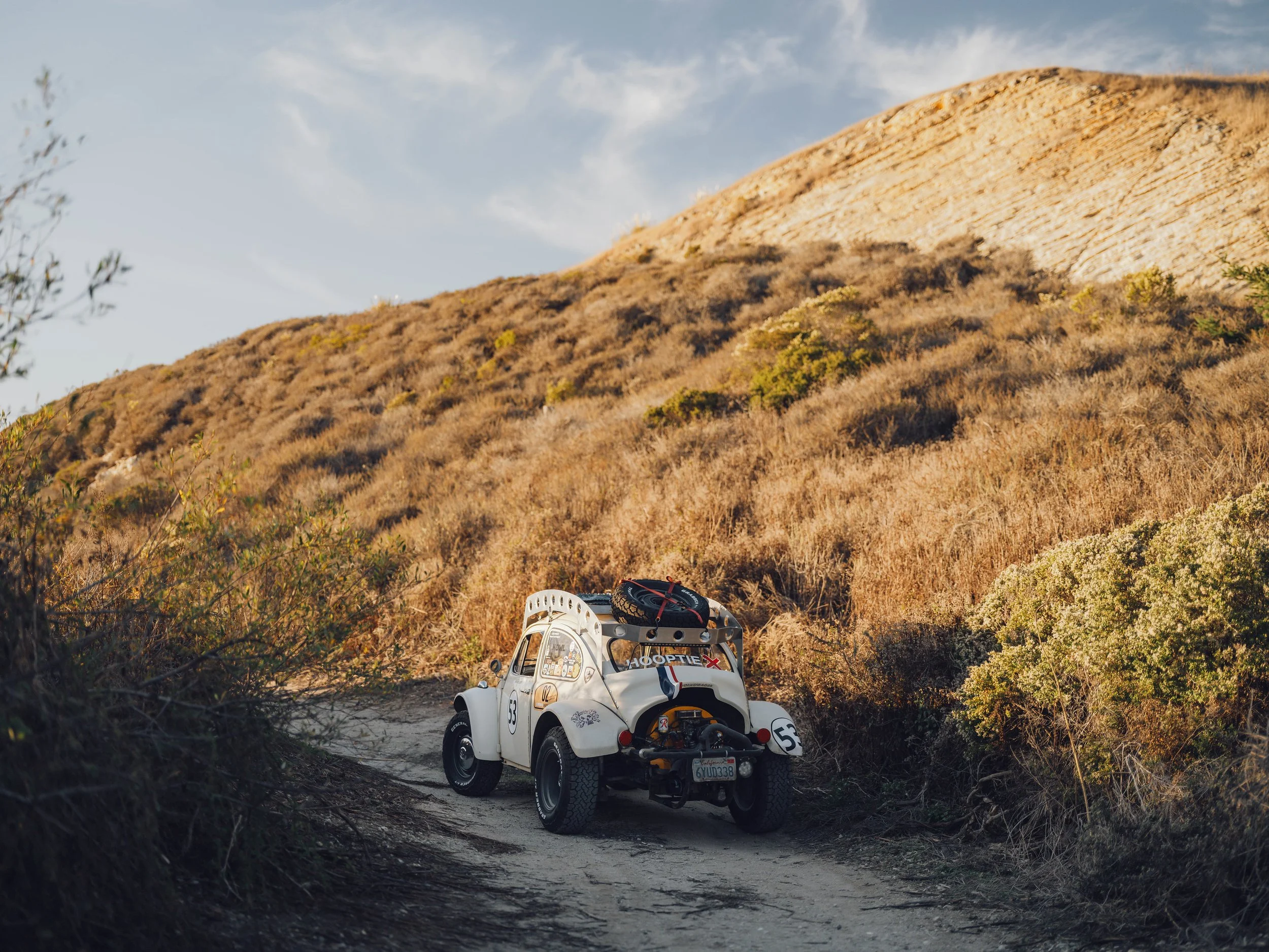 Vintage white dune buggy with racing number 53 on a dirt trail surrounded by dry grass and shrubs, with a mountainous landscape in the background under a partly cloudy sky.