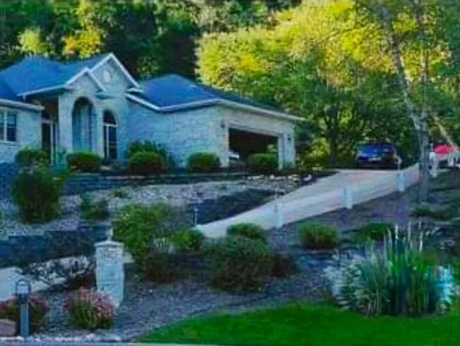A house with a gray stone exterior and a blue roof, situated on a hilly landscape with a driveway leading up to a garage. Trees and greenery surround the house.