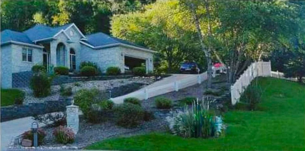 A house with a driveway leading up to a garage, surrounded by trees and landscaped yard with bushes and grass.