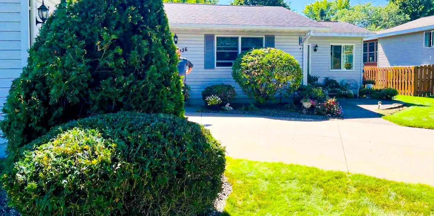 Front yard of a house with neatly trimmed bushes, a concrete driveway, green grass lawn, and a person standing near the house.