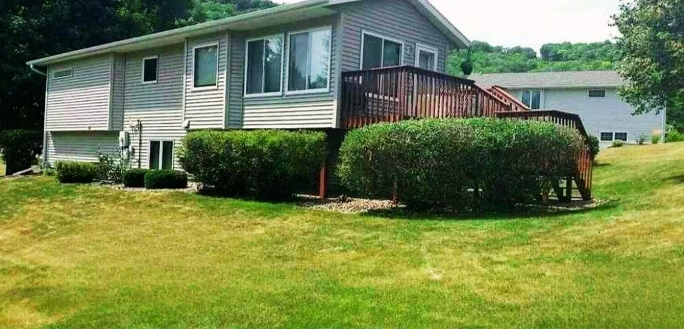 A house with a wooden deck and stairs facing a grassy yard, with bushes along the house and trees in the background.