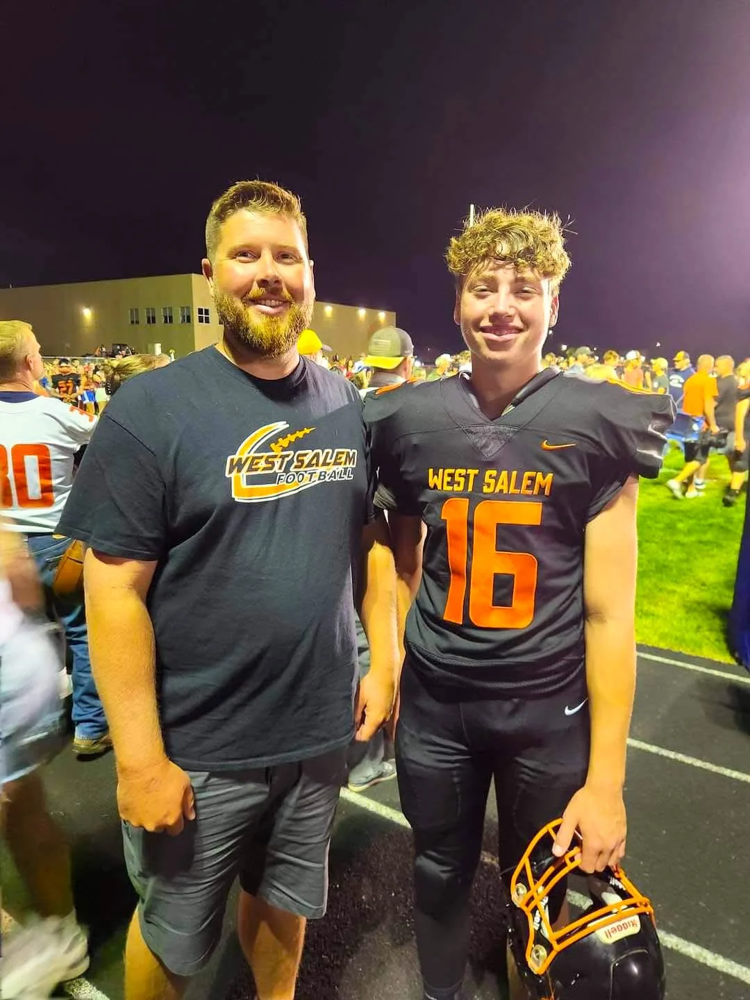 Two young men standing outdoors on a football field at night, smiling. The man on the left has a beard and is wearing a black West Salem football T-shirt, while the man on the right is in a black football uniform with the number 16 and holding an orange football helmet. Behind them, many other football players and spectators are visible.