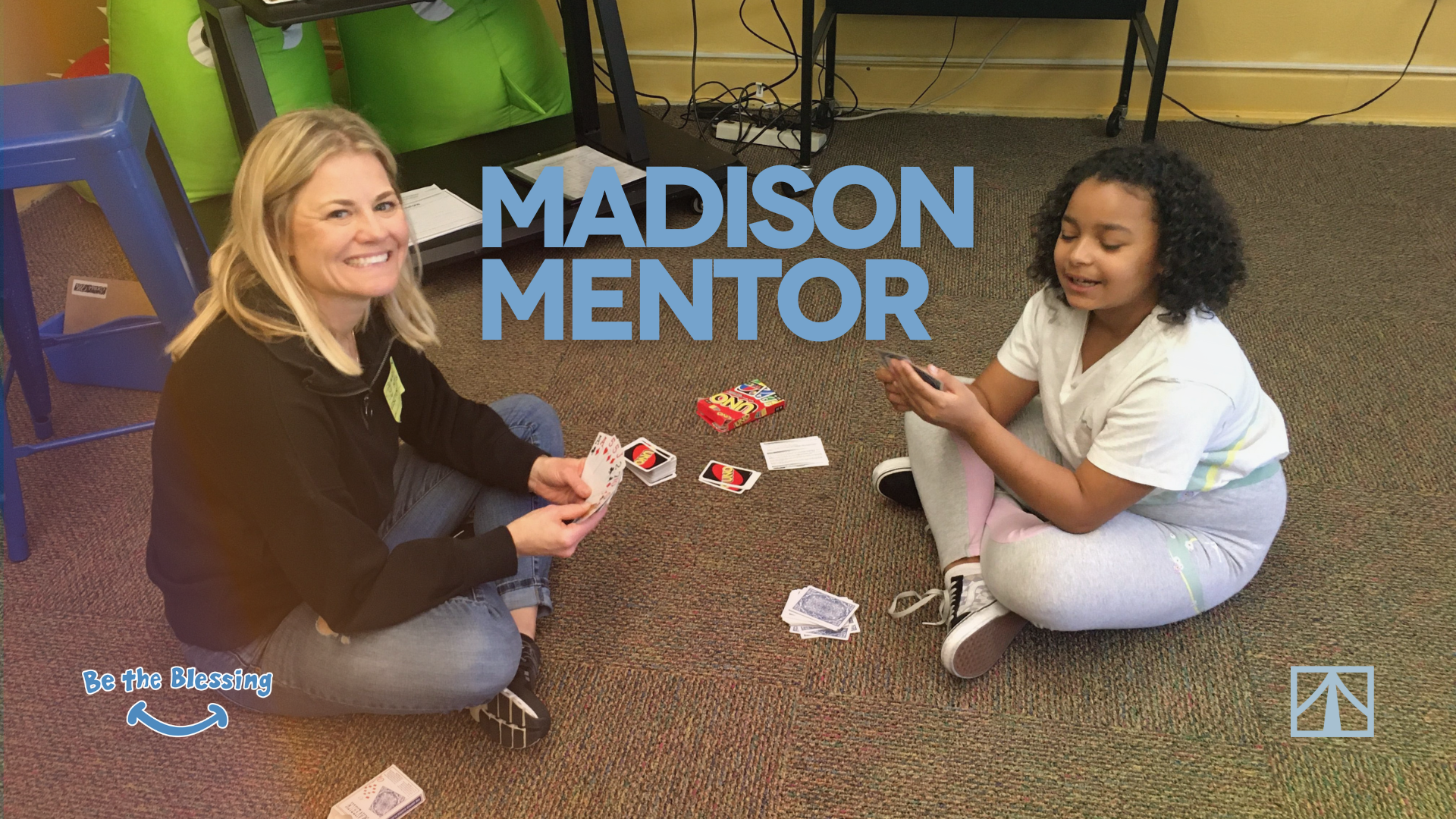 Two women sitting on the carpeted floor playing a card game. One woman has blonde hair, wearing a black jacket and jeans; the other with curly hair, wearing a white t-shirt and gray sweatpants. The scene includes a box of UNO cards, a deck of regular cards, and a small piece of paper. A blue chair and a black table with various cords are in the background, along with colorful cushions and a poster that says "Be the Blessing." The overlay text reads "Madison Mentor."