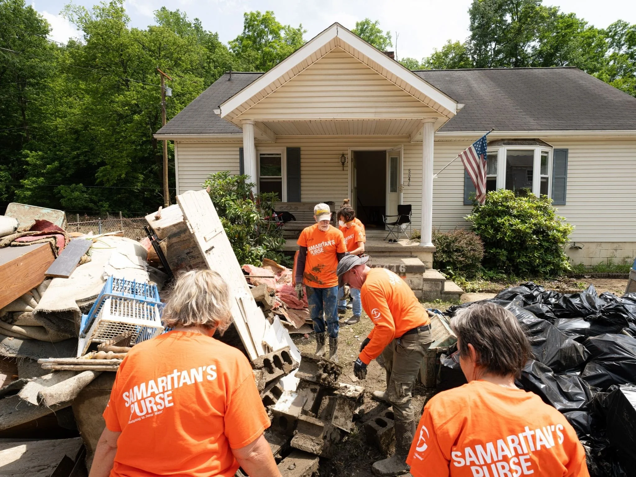 Group of volunteers in Orange shirts removing debris from a house front yard after a disaster, with a house and greenery in the background.