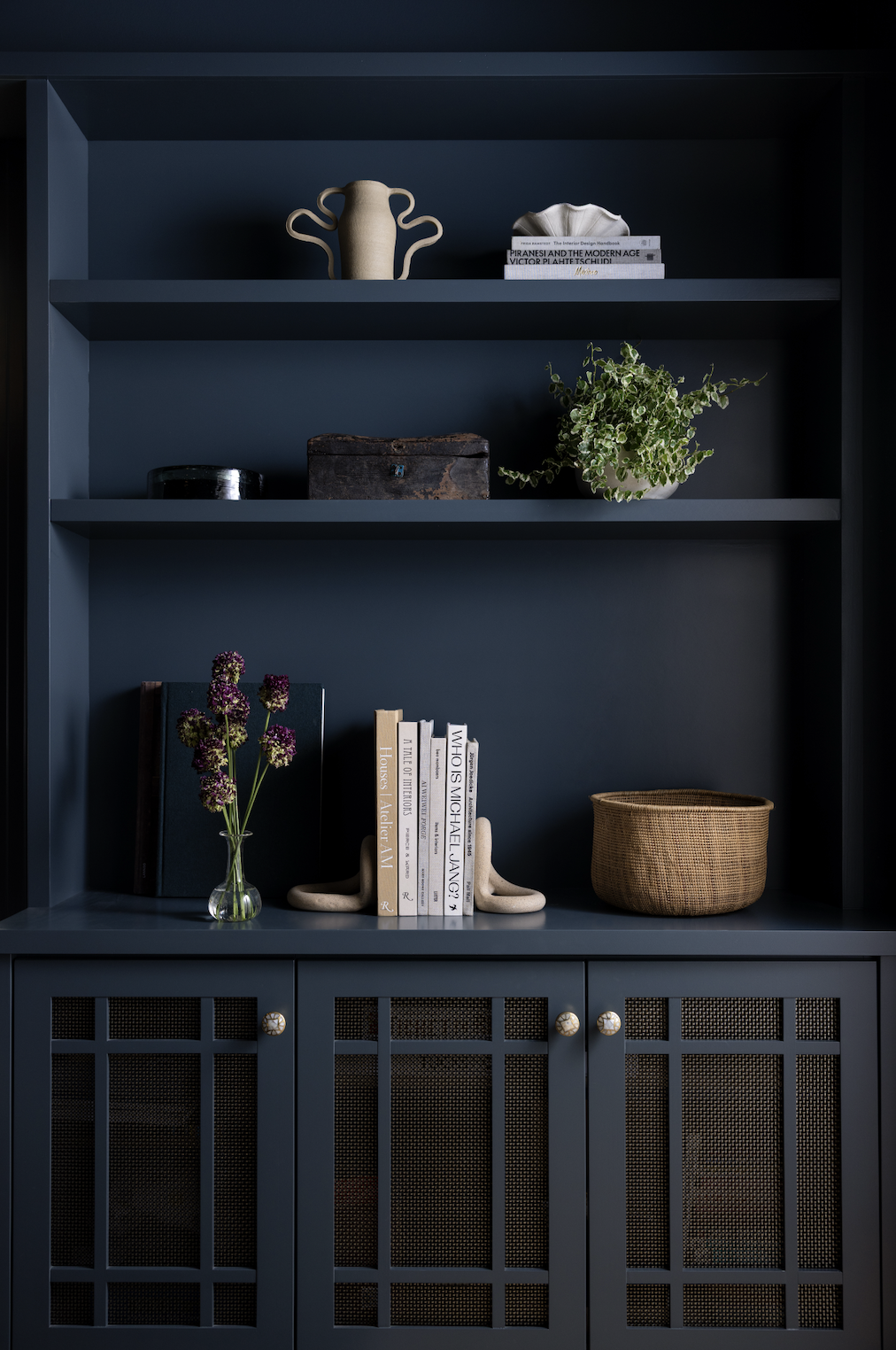 Built-in navy shelving styled with decorative ceramics, design books, greenery, and woven storage baskets.