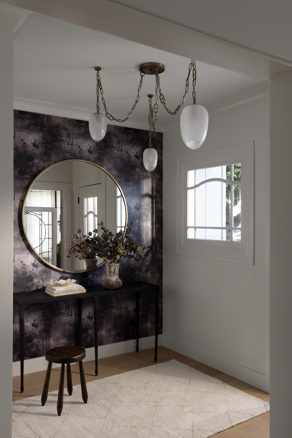 Entryway featuring dramatic botanical wallpaper, a round brass-framed mirror, a slim black console, and sculptural pendant lighting.
