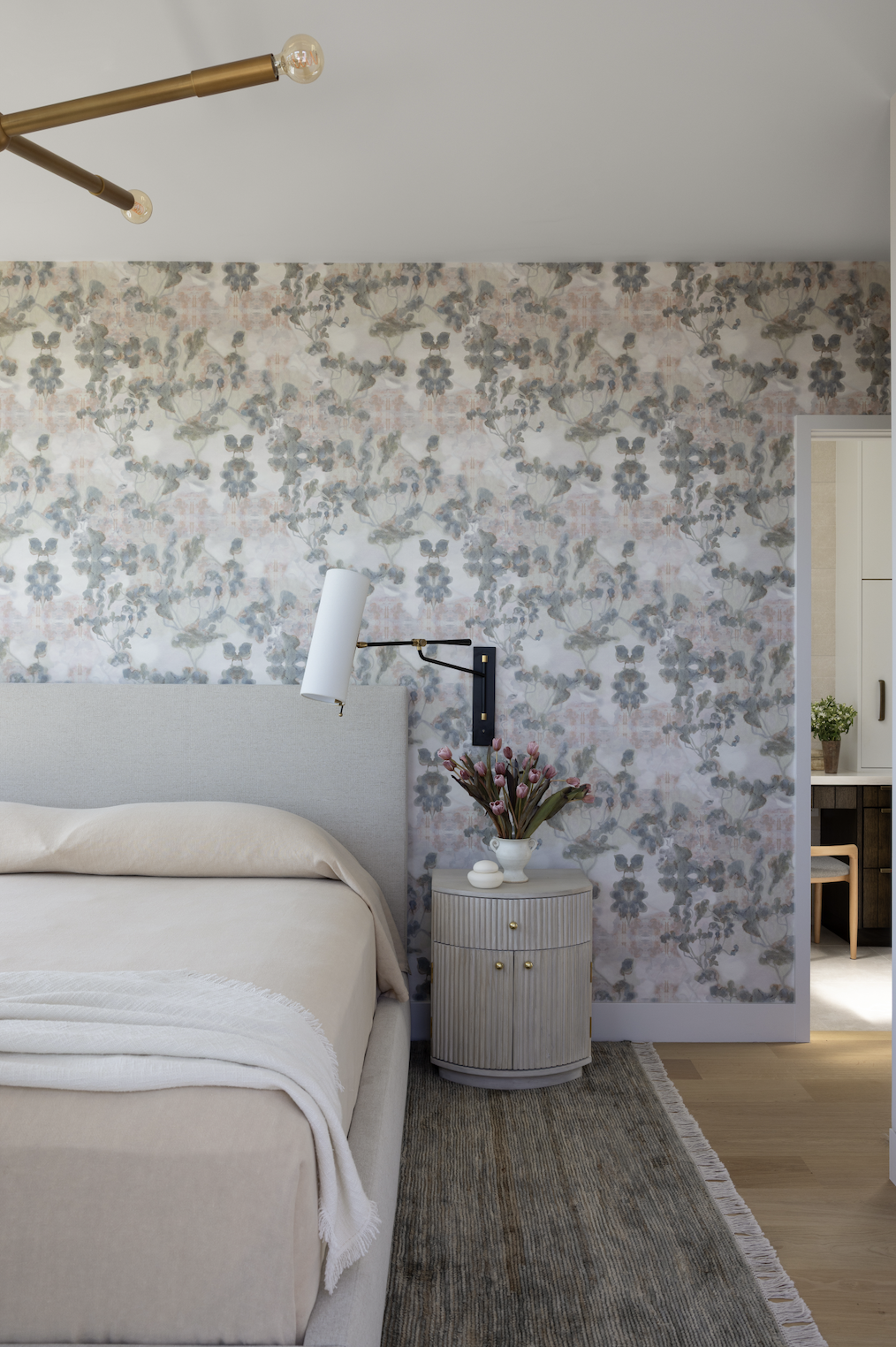 Bedroom featuring soft blue and blush floral wallpaper, a cream upholstered bed, brass lighting, and a fluted bedside table styled with fresh flowers.