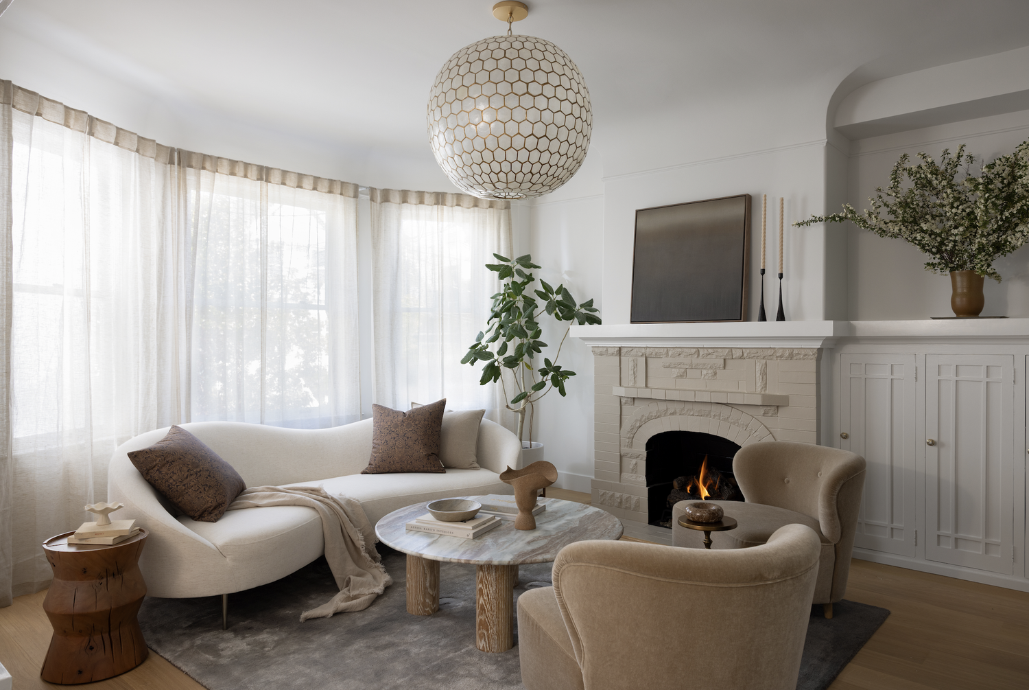 Light-filled living room with a curved cream sofa, sculptural marble coffee table, textured fireplace, and warm neutral furnishings.