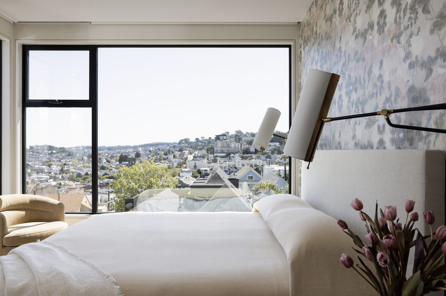 Bedroom with soft floral wallpaper, an upholstered cream headboard, brass lighting, and muted pink accents.