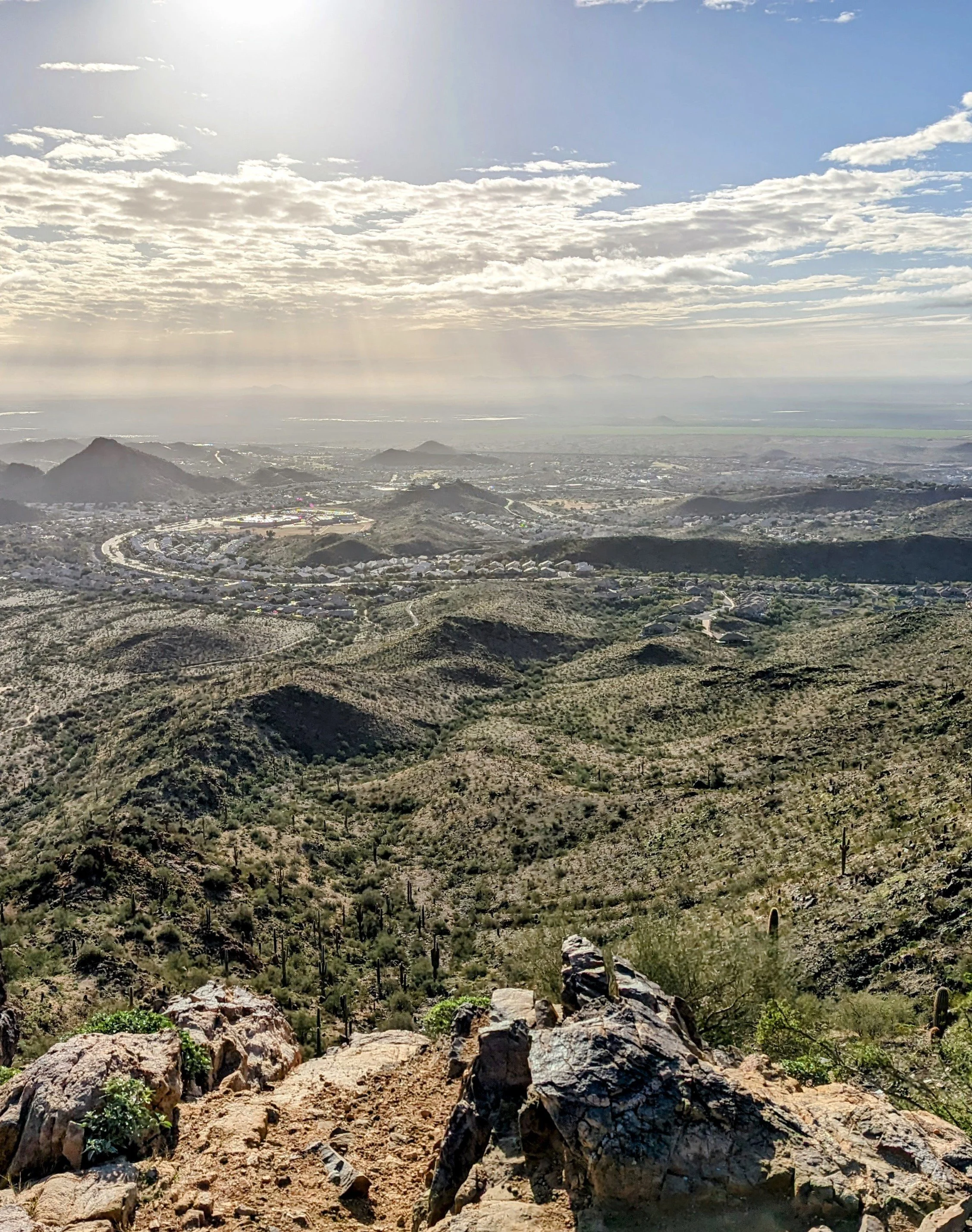 A panoramic view from a mountain overlook showing a desert landscape with rocky terrain, cacti, rolling hills, and distant city or residential areas under a cloudy sky with sunlight breaking through.