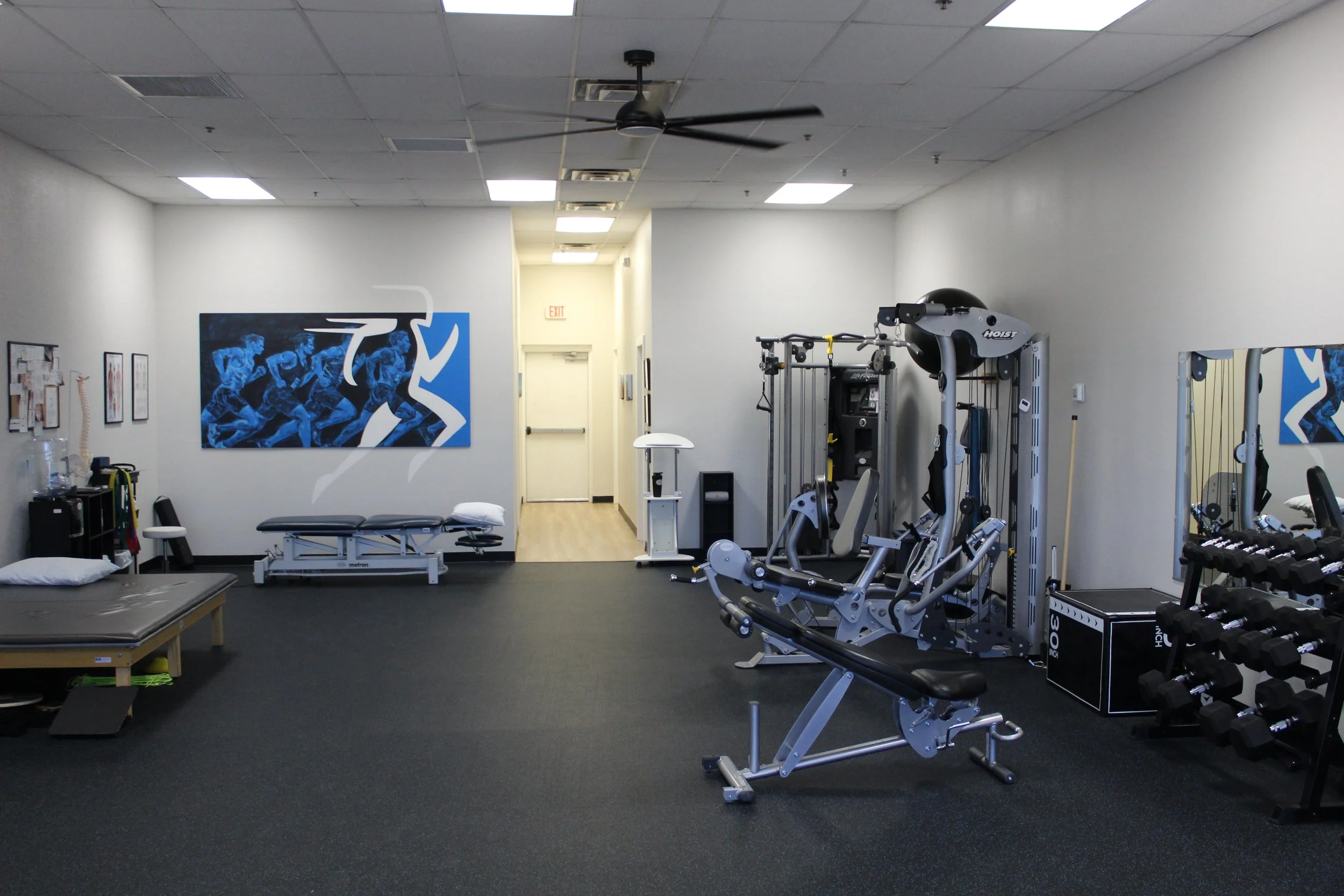 A fitness gym with workout equipment including a seated row machine, dumbbell rack, therapy table, and a poster of running athletes on the wall.