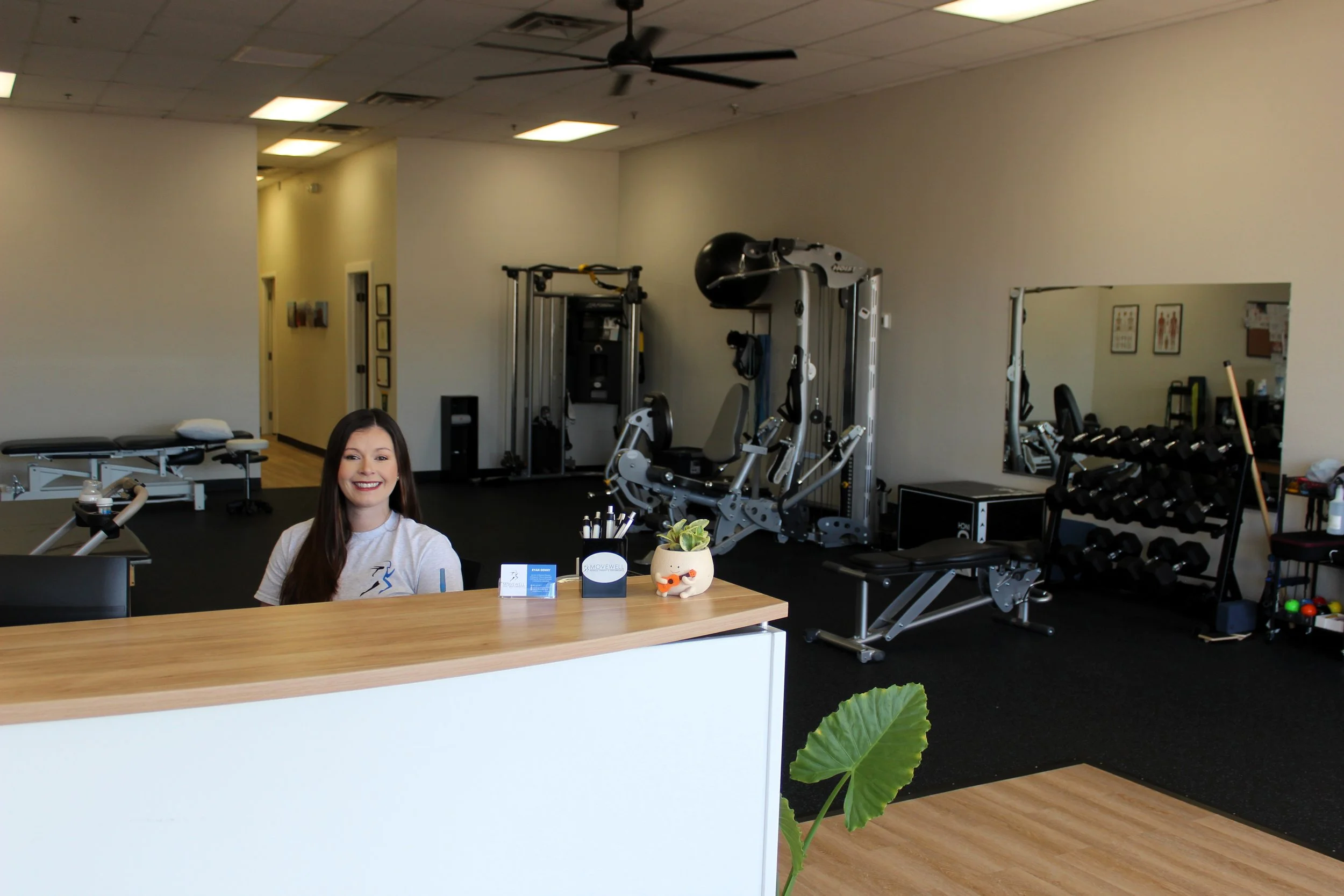Reception area of a physical therapy clinic with a smiling woman behind the counter, exercise equipment, and fitness machines in the background.