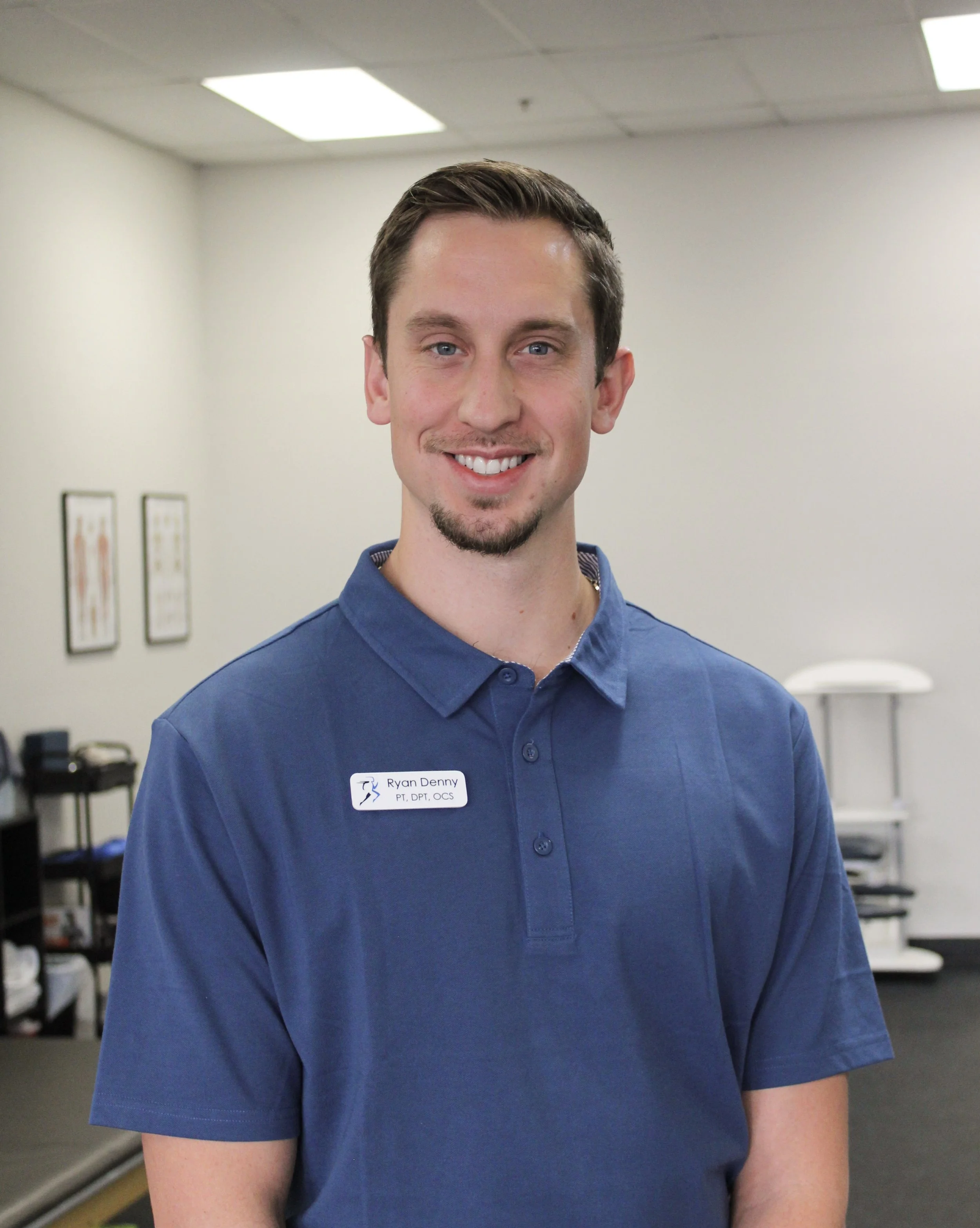 A young man with short brown hair, a goatee, and blue eyes, smiling and wearing a blue polo shirt with a name tag that reads 'Ryan Denny PT, DPT, OCS'. He is standing in a clinical or office setting with framed pictures on the wall and medical equipment in the background.