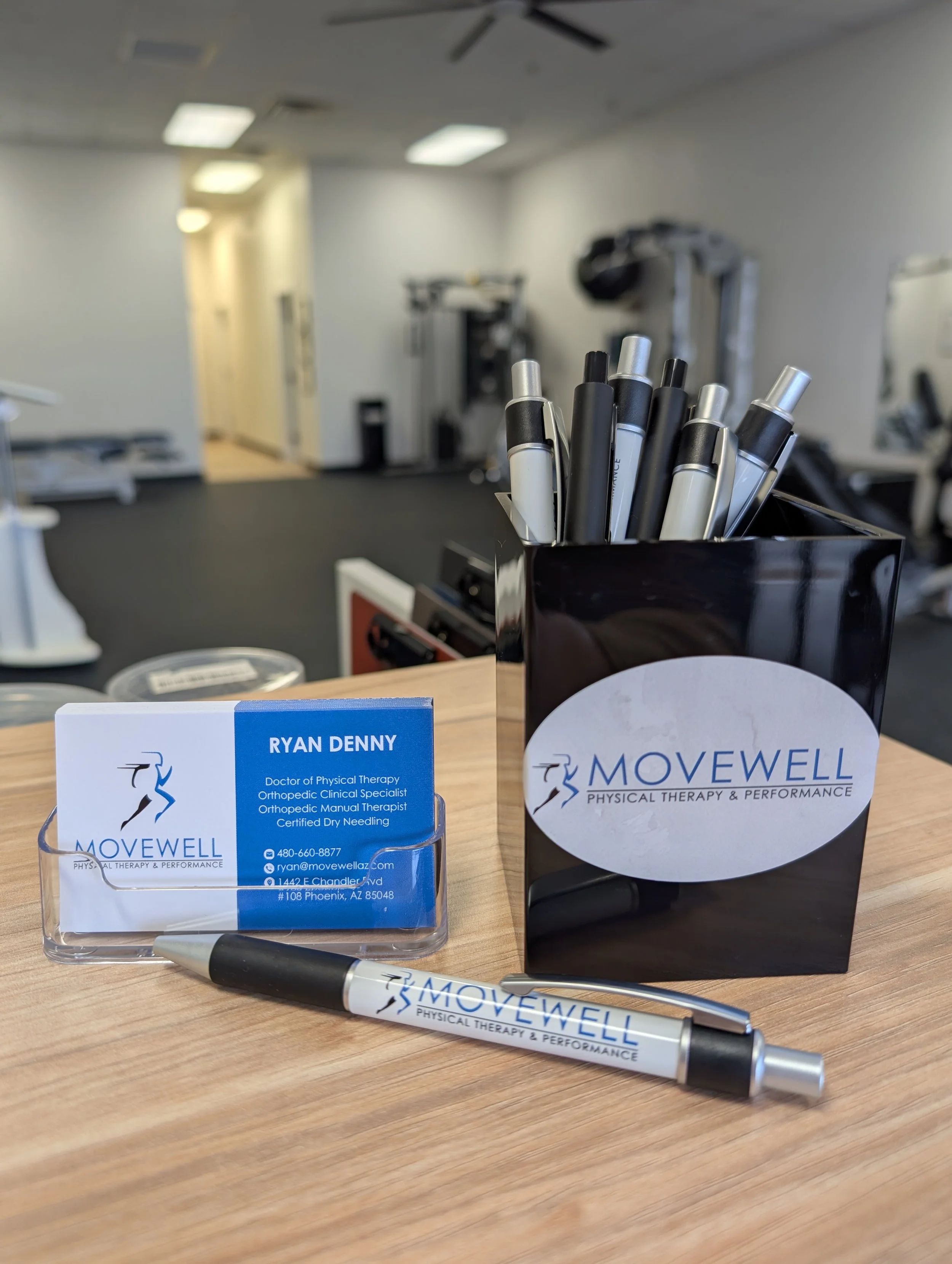 Business card, pen, and black pen holder with multiple markers on a wooden desk in a physical therapy clinic.