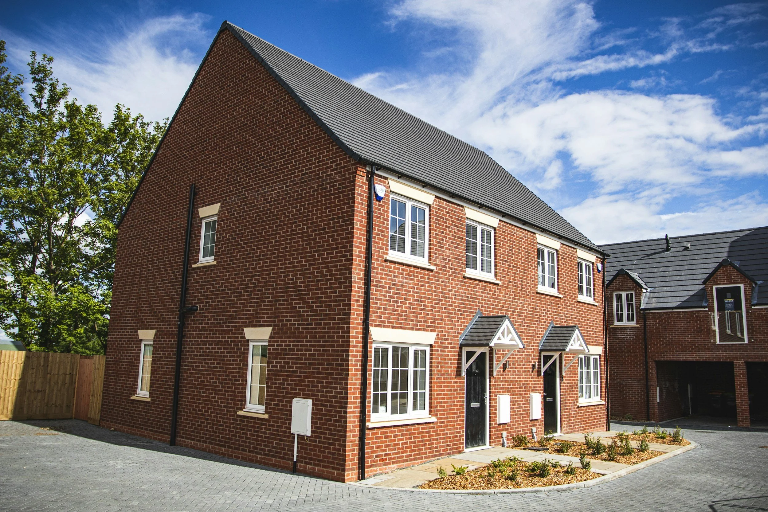 Newly built red brick houses with black doors and small front gardens under a blue sky with clouds.