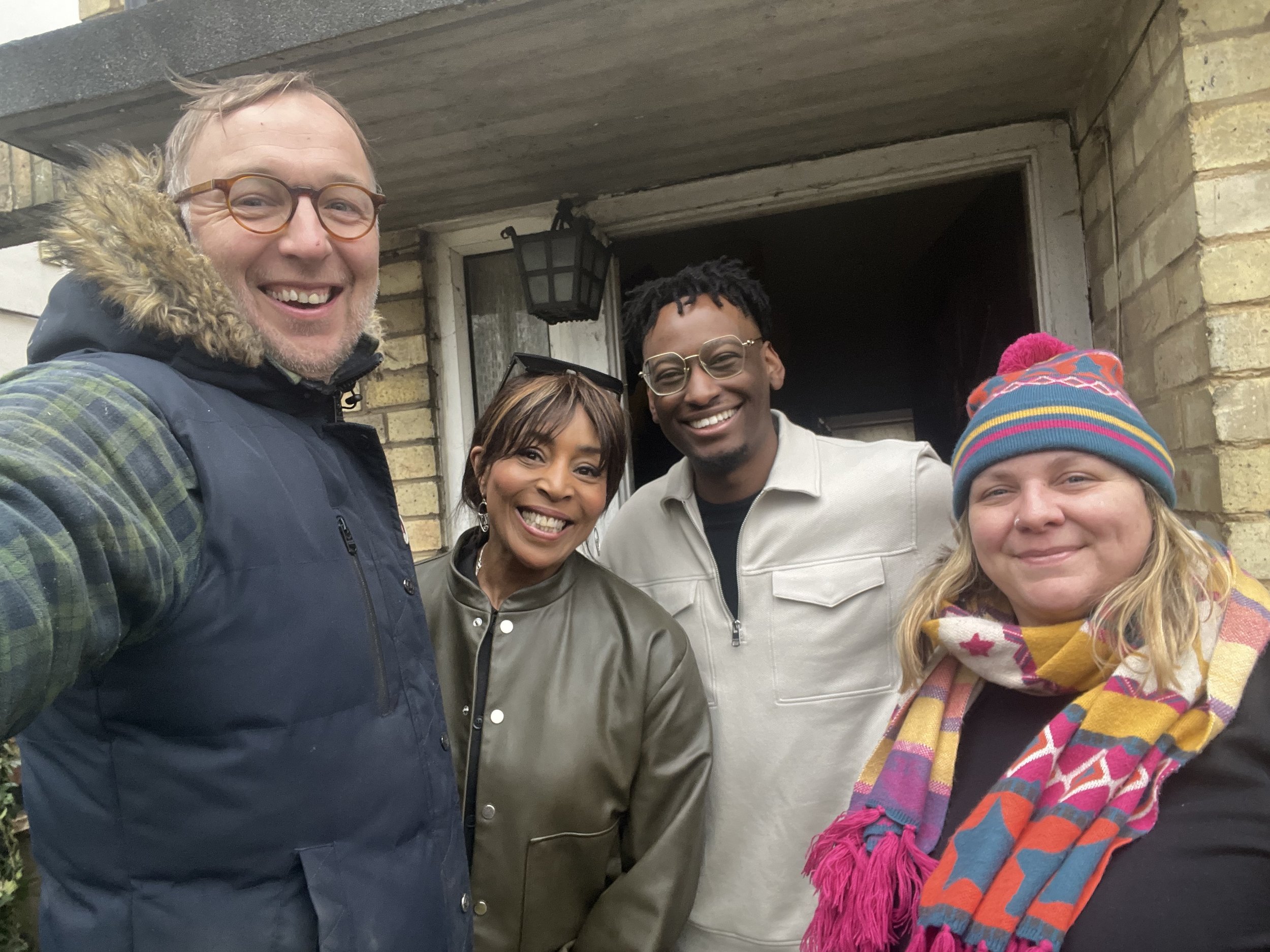 Four smiling people standing outside a house, taking a selfie. The person on the front left wears glasses and a vest with a fur-lined hood. The woman in the center wears earrings and a green jacket. The man next to her has glasses and a light-colored jacket. The woman on the right wears a colorful hat and scarf.