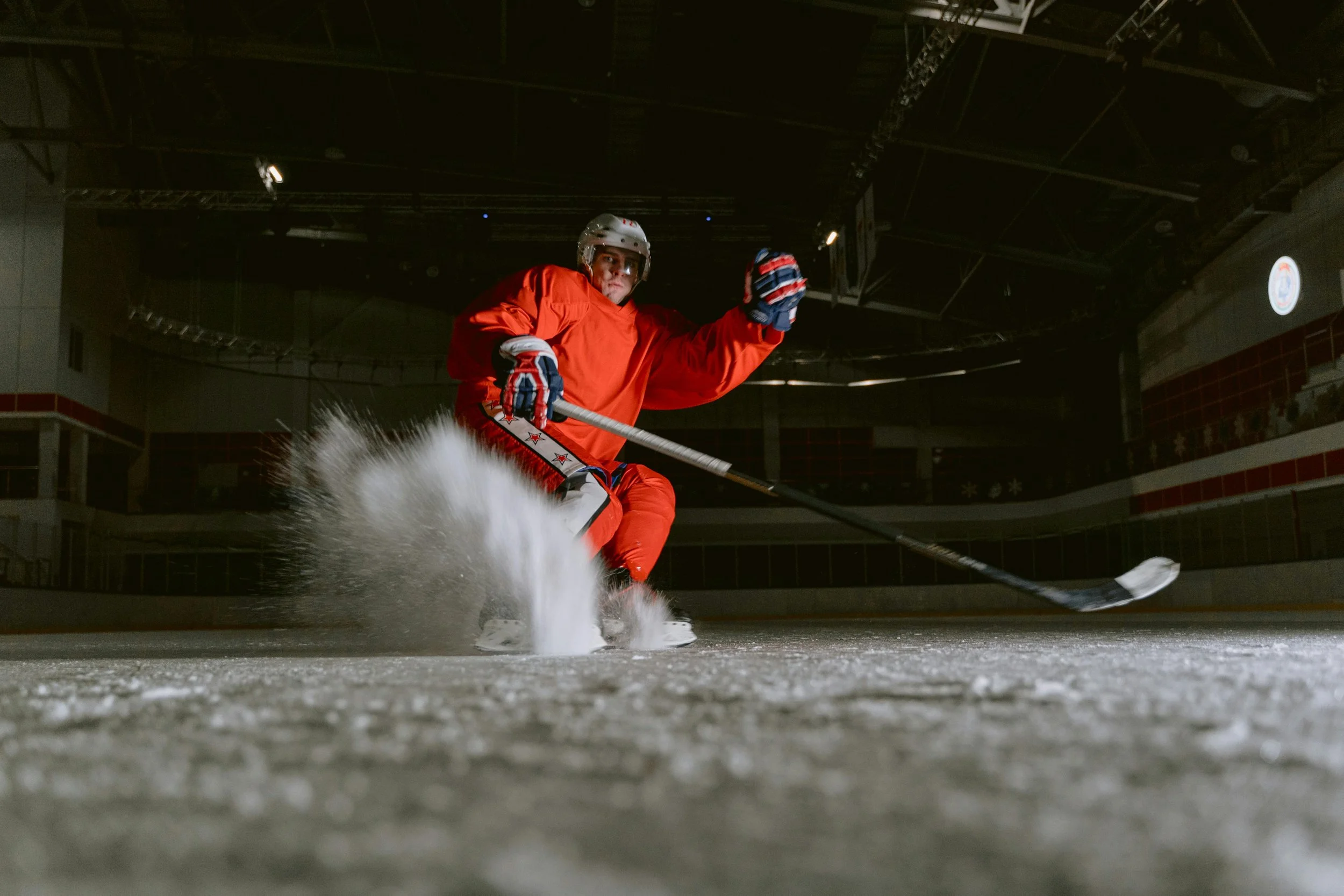 A hockey player wearing a red uniform and helmet is skating on an indoor ice rink, with snow spray behind him as he maneuvers with a hockey stick.
