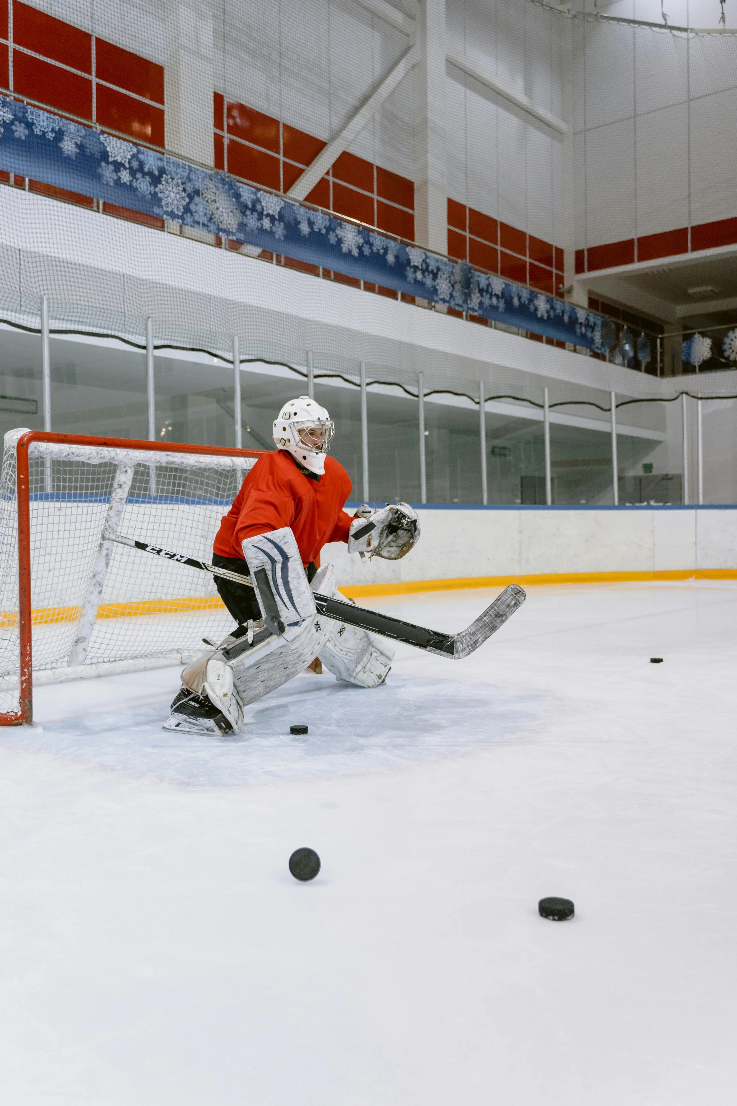 A hockey goalie on the ice during practice or game, wearing red jersey, white helmet, and protective gear, positioned in front of the goal with hockey pucks scattered on the ice.