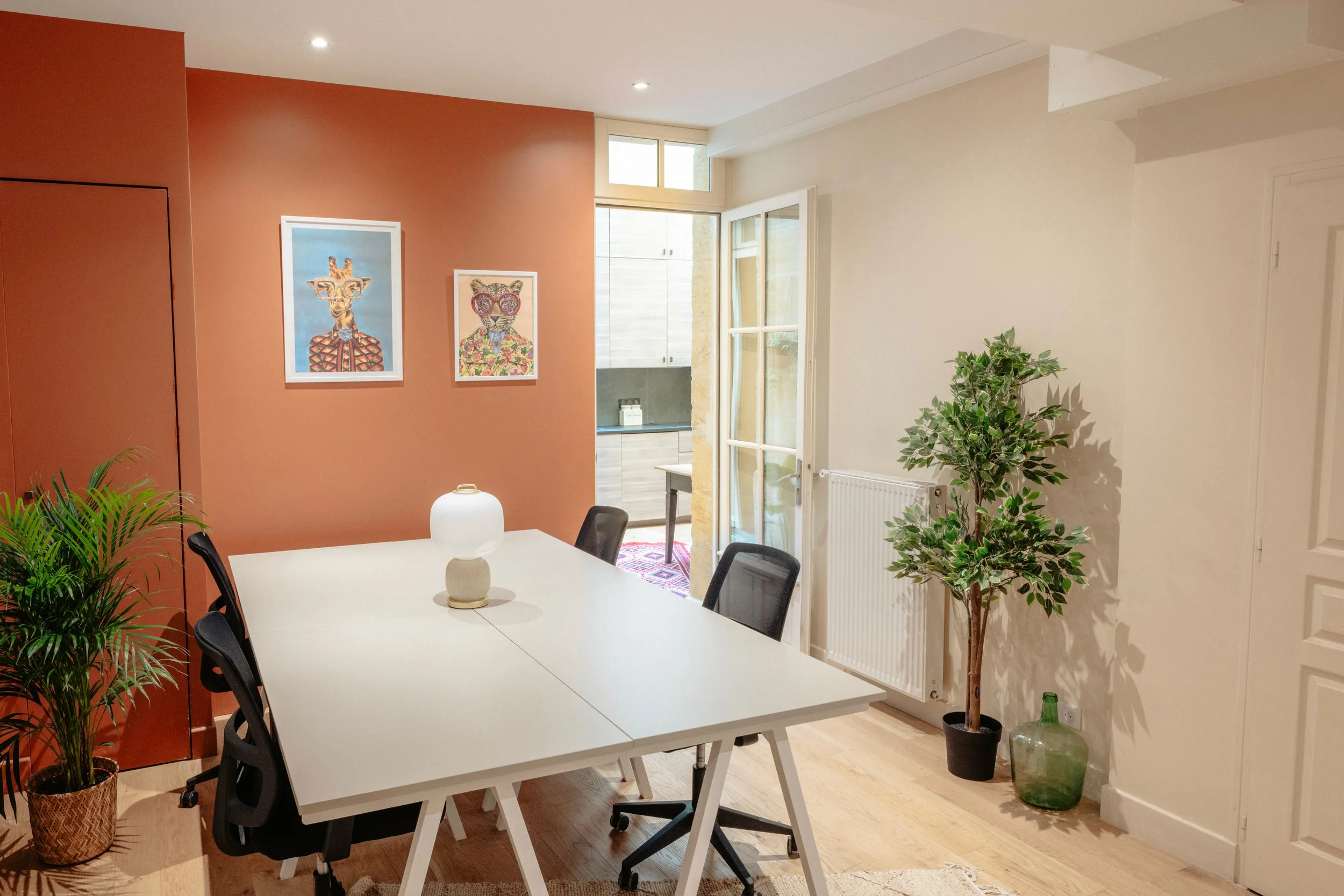 Salle à manger moderne avec table blanche, chaises noires, mur terracotta avec deux œuvres d'art, plantes vertes, porte-fenêtre donnant sur une cuisine.