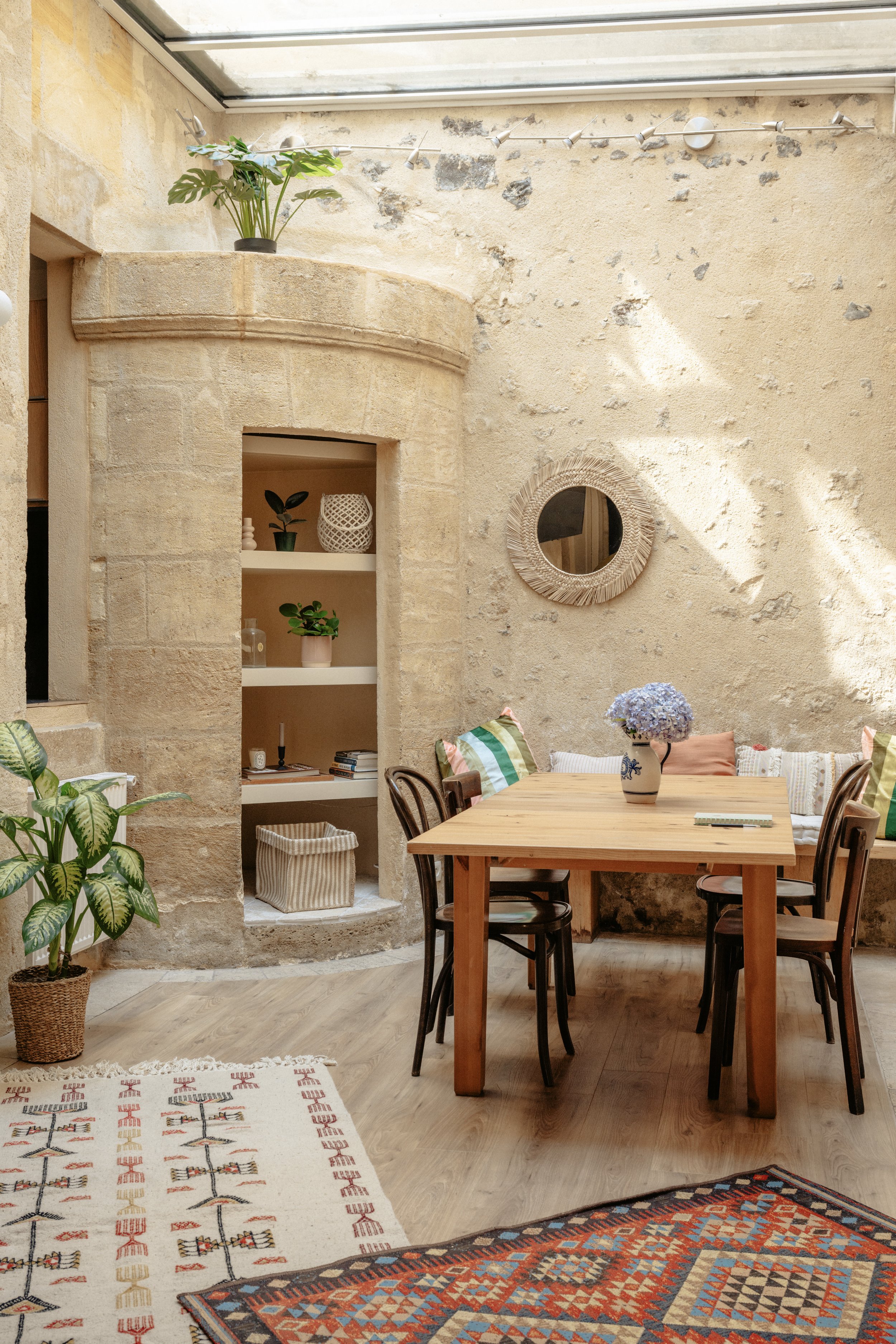 Intérieur d'une salle à manger avec un mur en pierre, une table en bois avec des chaises et des coussins colorés, une plante verte en pot, une étagère encastrée avec des décorations, et un miroir rond en rotin, très beau puit de lumière naturelle.
