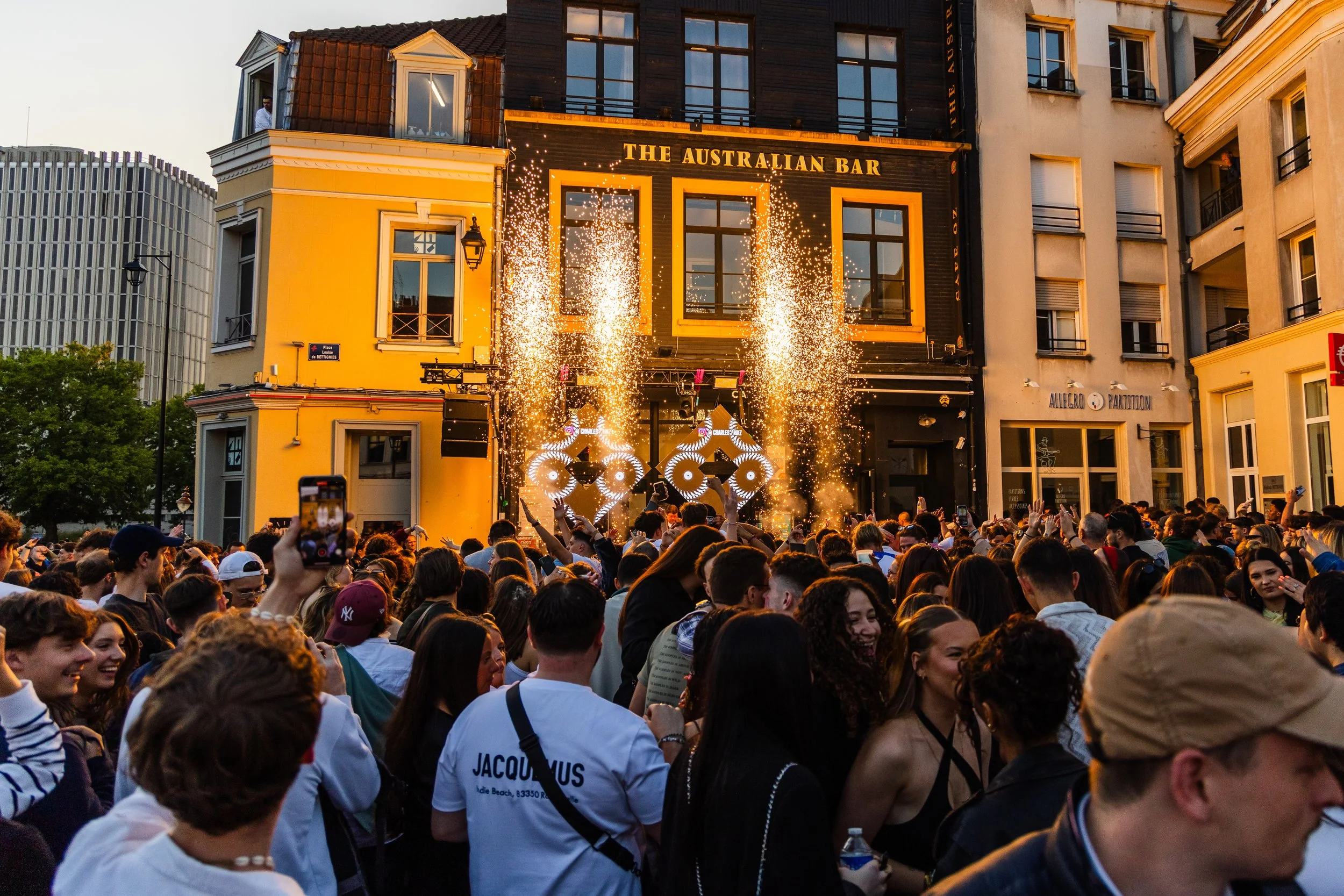 Fête en plein air avec un grand rassemblement de personnes devant un bar australien, avec des feux d'artifice et des performances de DJ sur scène, en soirée.