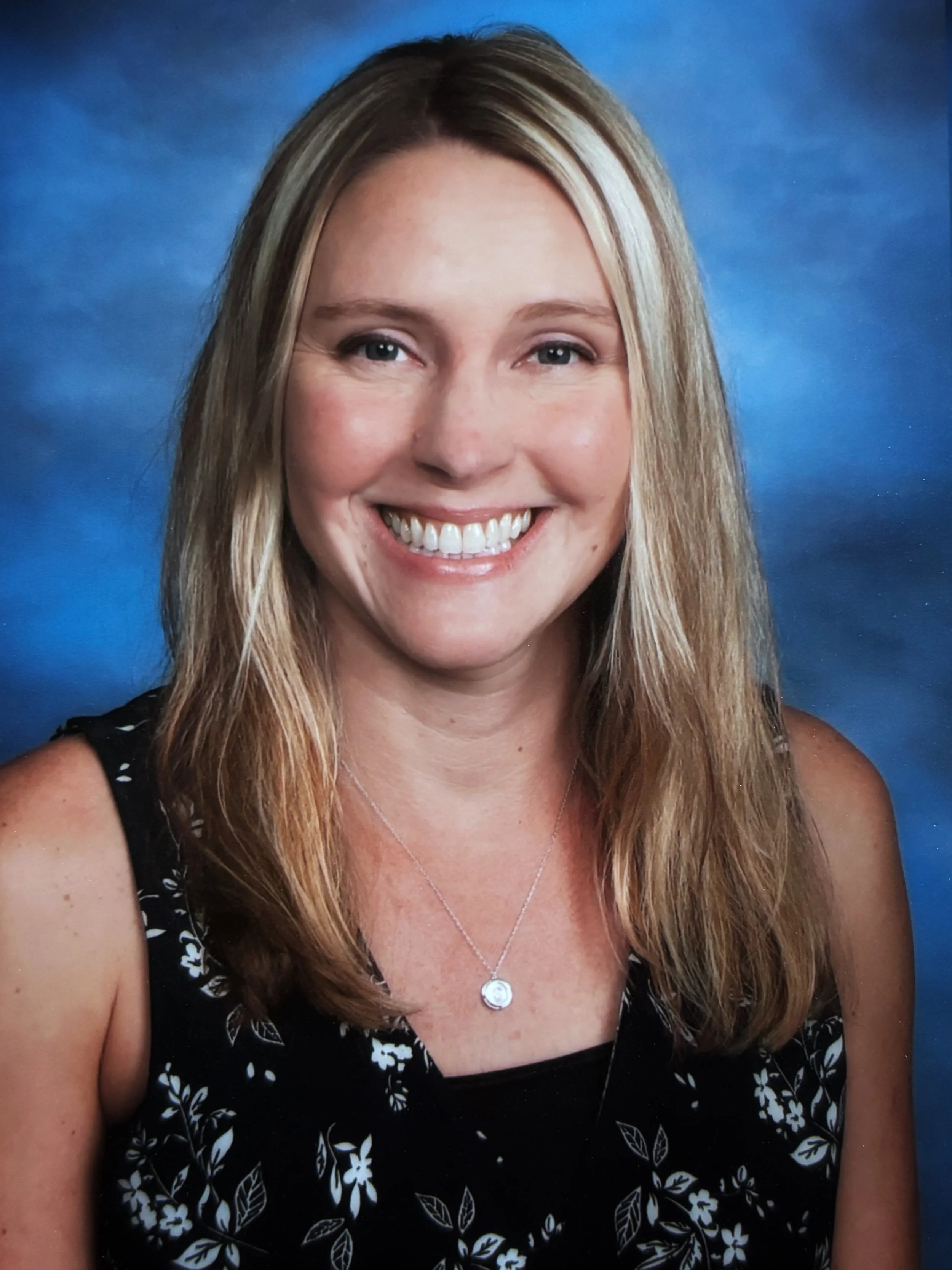 Close-up of a woman with blond hair, glasses, and a blue top, smiling outdoors.