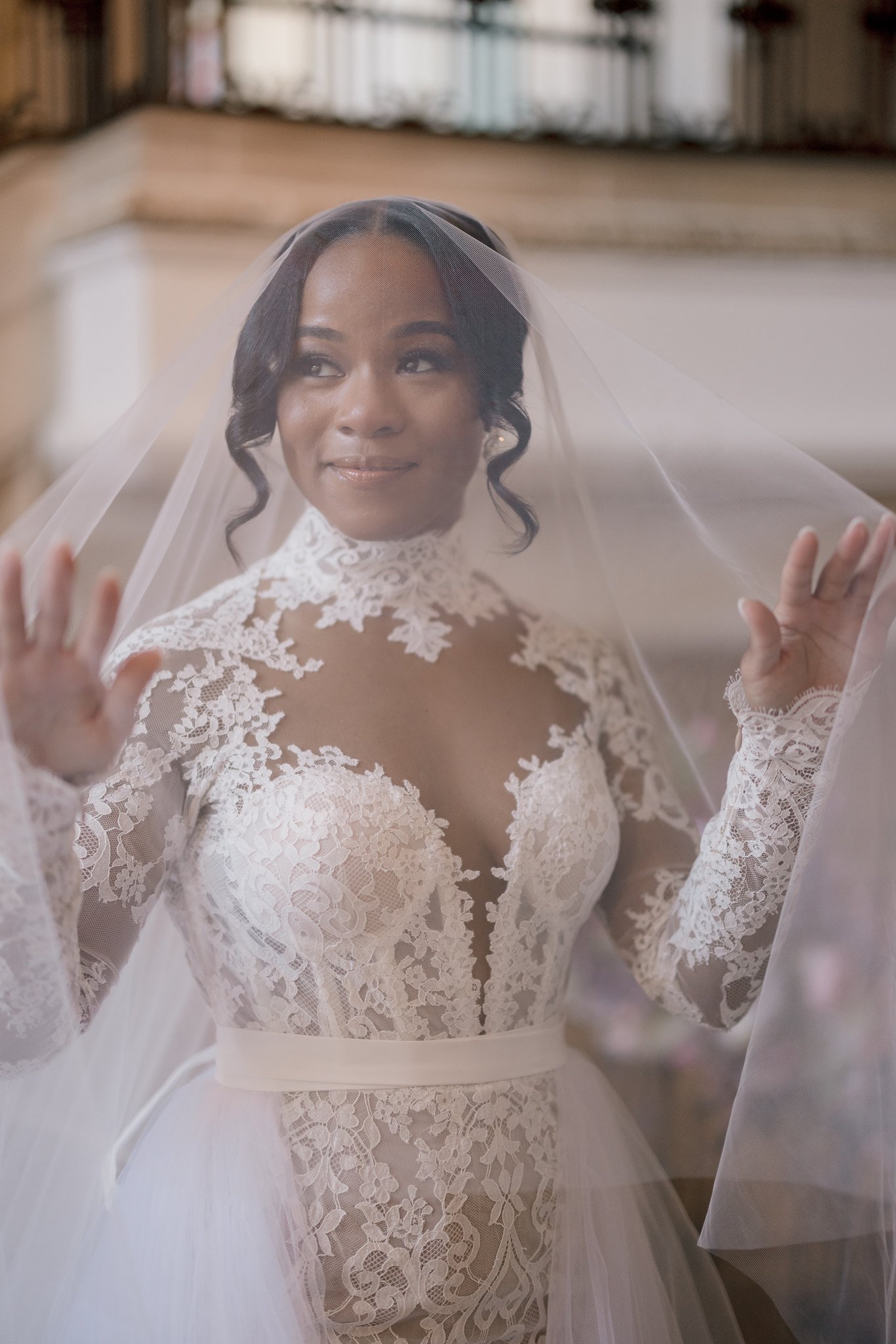 A bride dressed in a lace wedding gown with a sheer veil, standing indoors with a gentle smile.