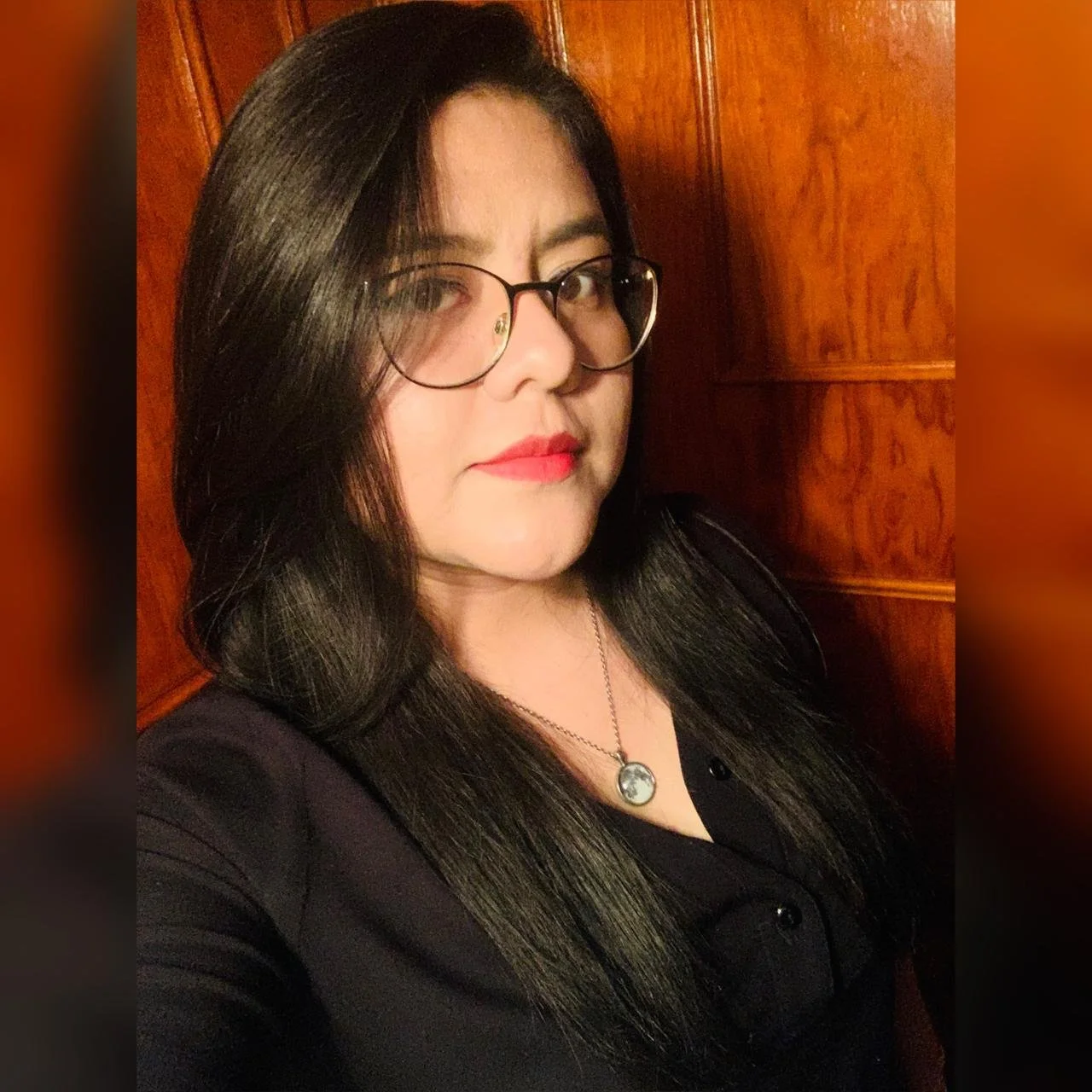 Retrato de una mujer con gafas, cabello largo y oscuro, maquillada con labial rojo, vestida con blusa negra, con fondo de madera.