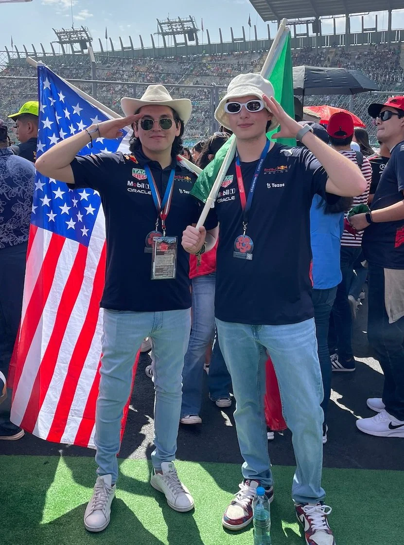 Two individuals at a racing event, saluting while holding flags, wearing sunglasses and casual clothing, with a crowd and a race track in the background.