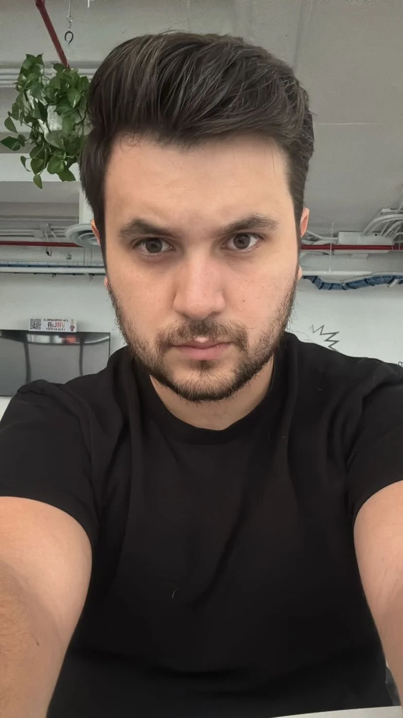 Joven con cabello oscuro y barba, tomando selfie en lo que parece ser una oficina o espacio de trabajo.