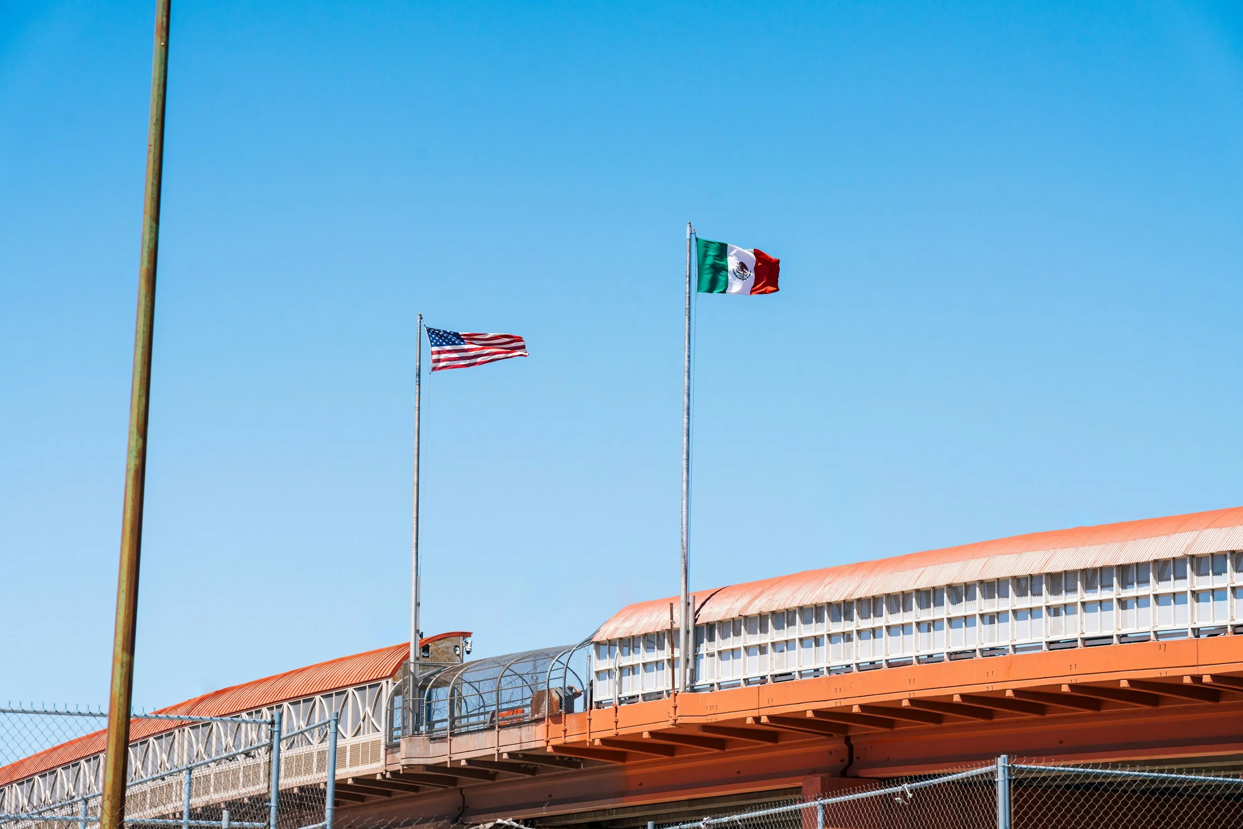 Banderas de Estados Unidos y México ondeando en un cielo azul, con un edificio con estructura metálica y ventanas en el fondo.