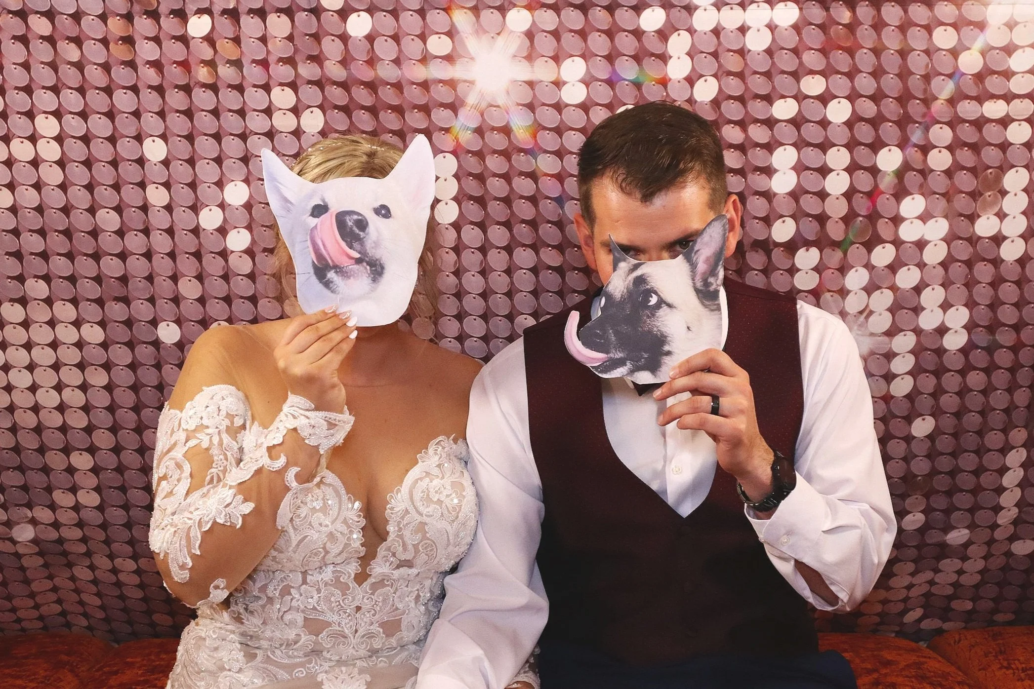 A bride and groom holding paper masks of dogs over their faces, sitting against a pink, sequined background.