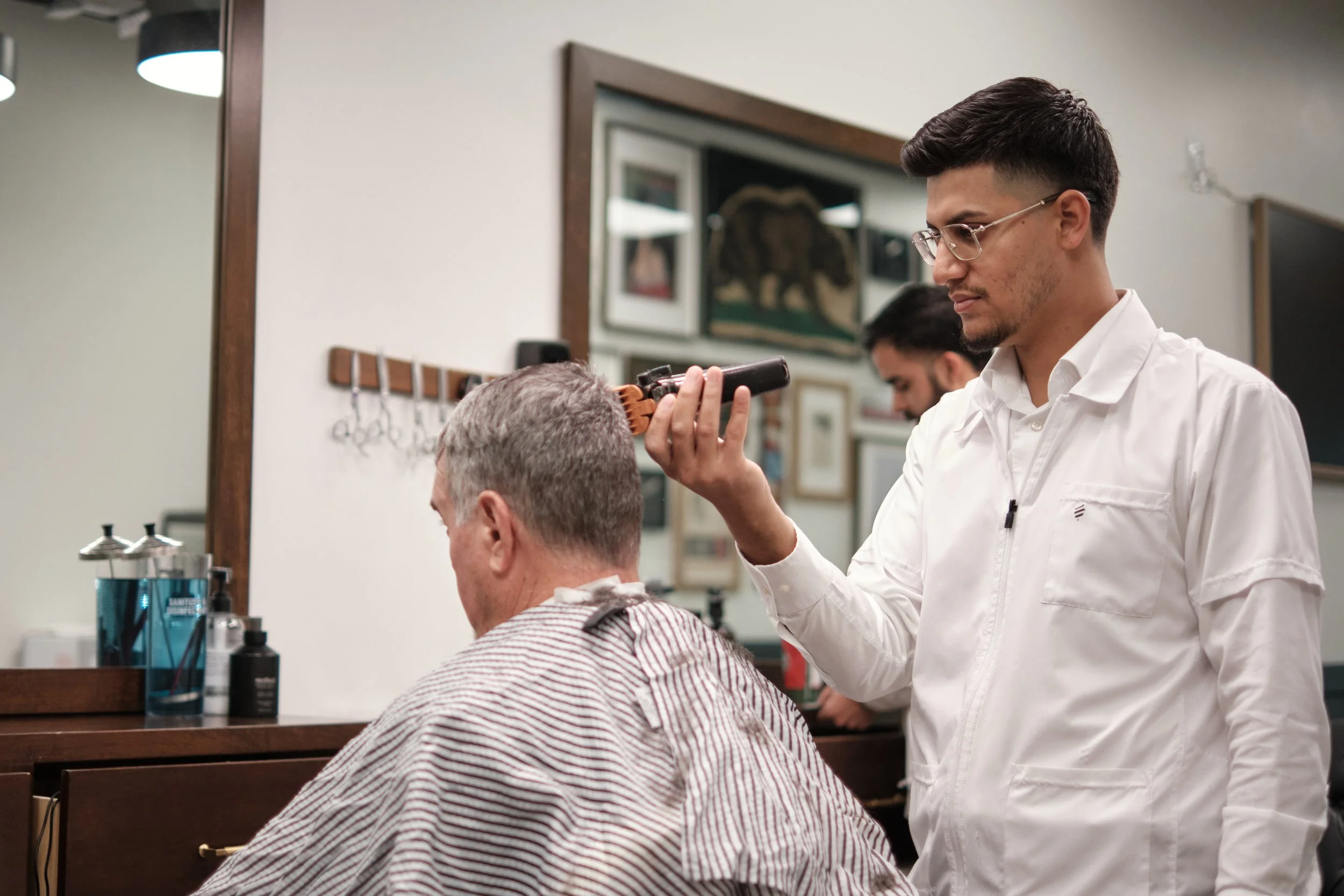A young man wearing a white barber coat and glasses gives a haircut to an older man with gray hair, sitting in a barber shop chair.