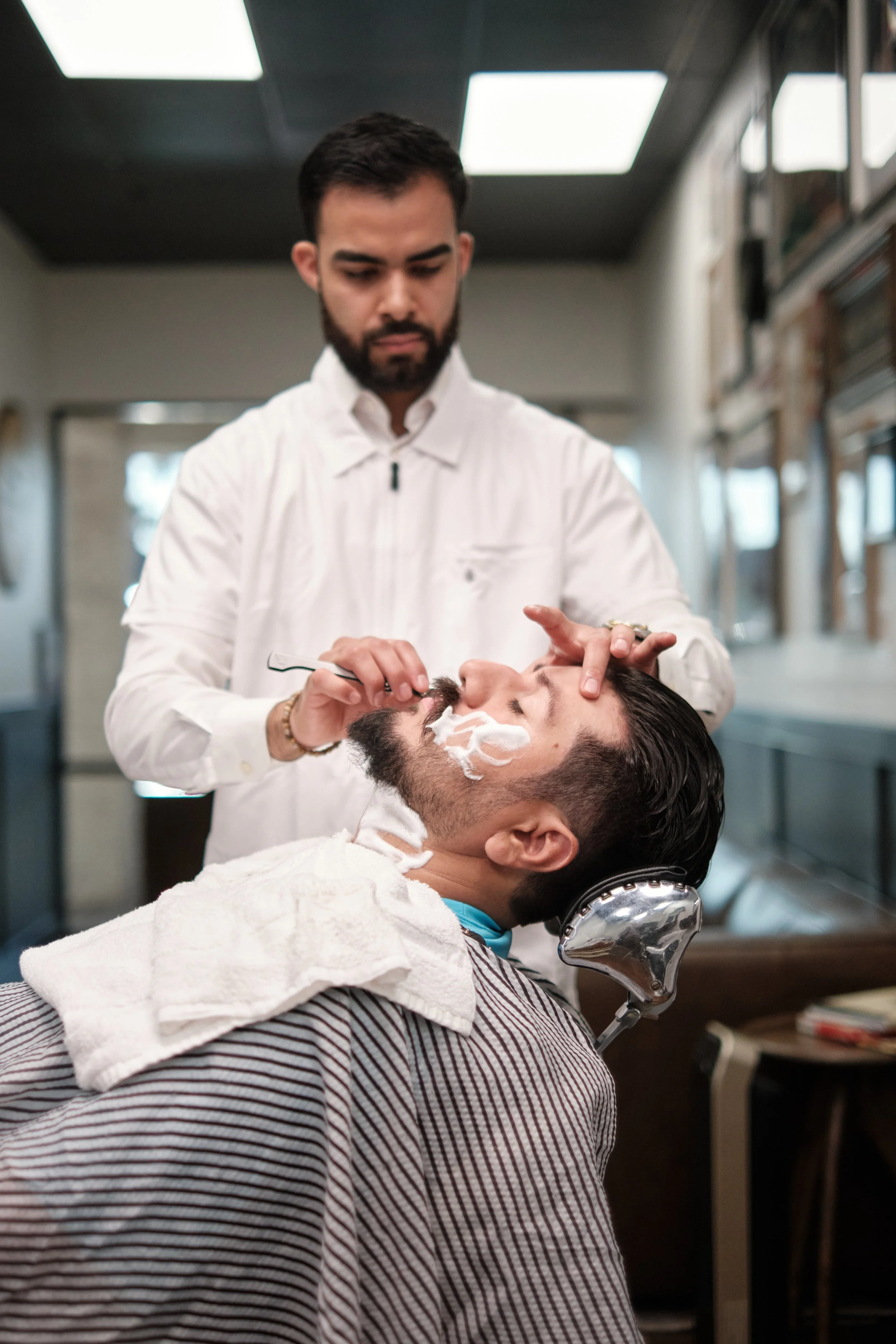 A man getting a shave at a barbershop. The barber is applying shaving cream to the man's face while he reclines in a barber chair.